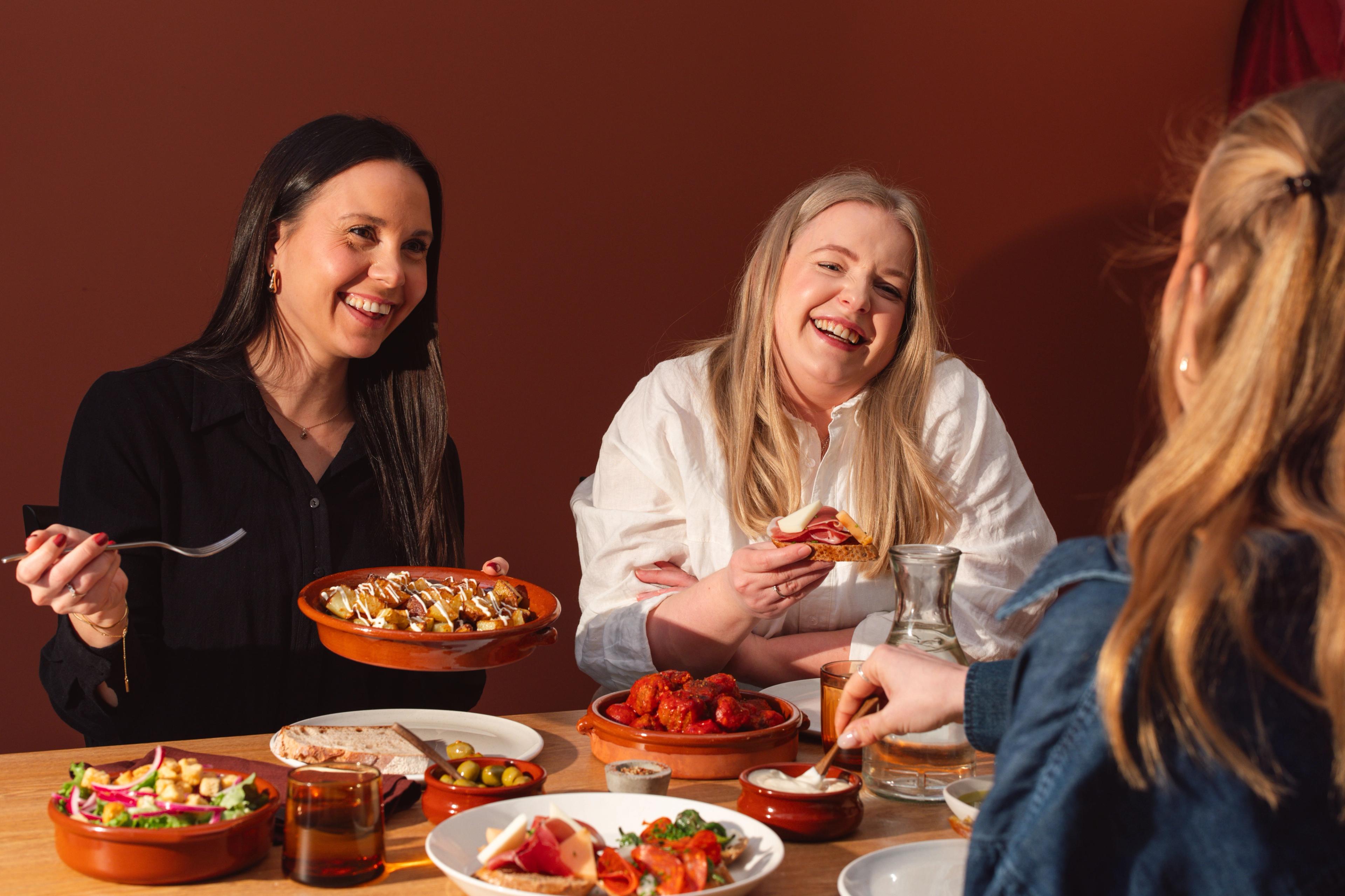 To smilende kvinner sitter ved et bord dekket med skåler og fat med spanske retter