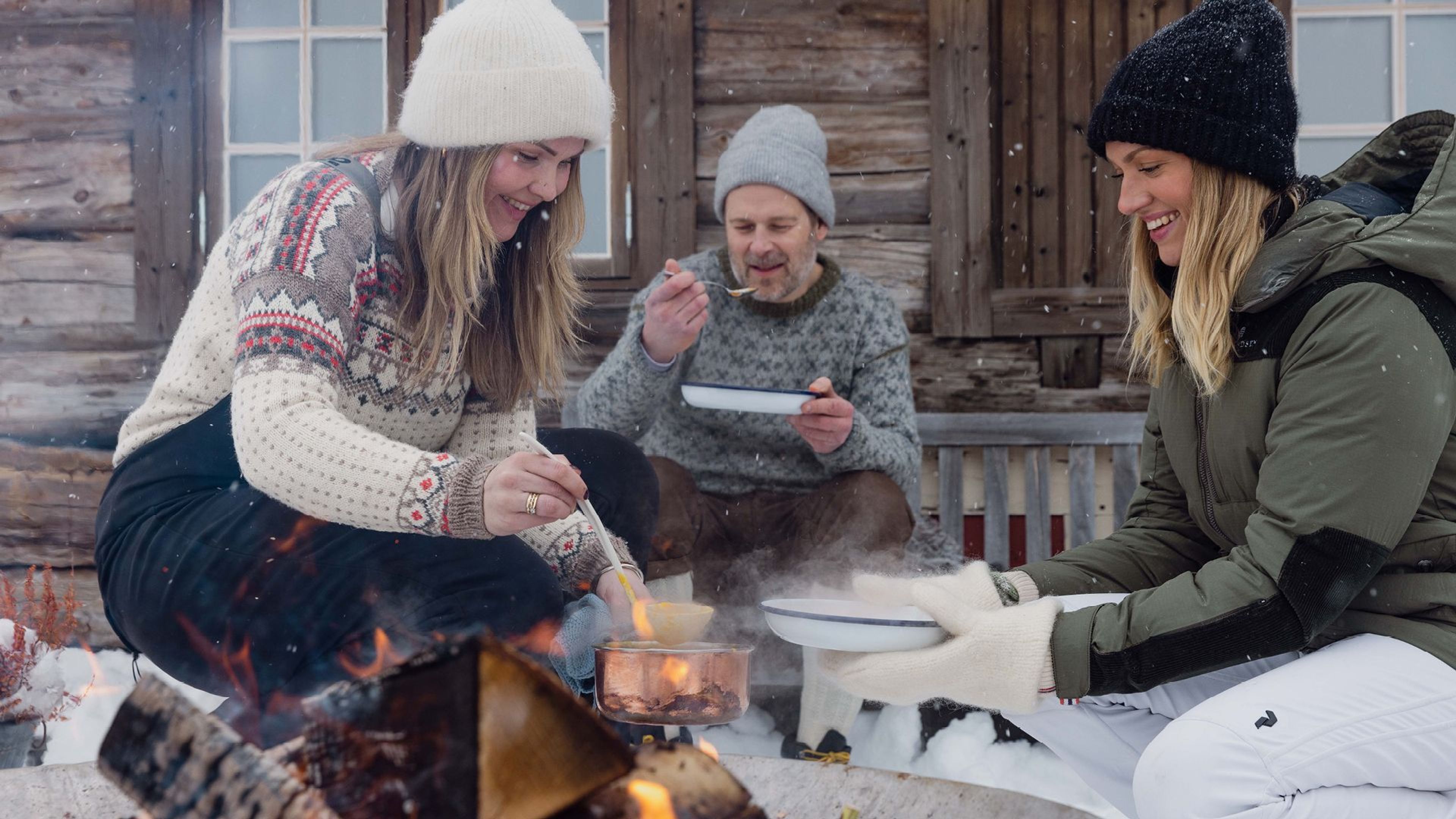 Glade personer lager mat rundt et leirbål utenfor en hytte i snøen