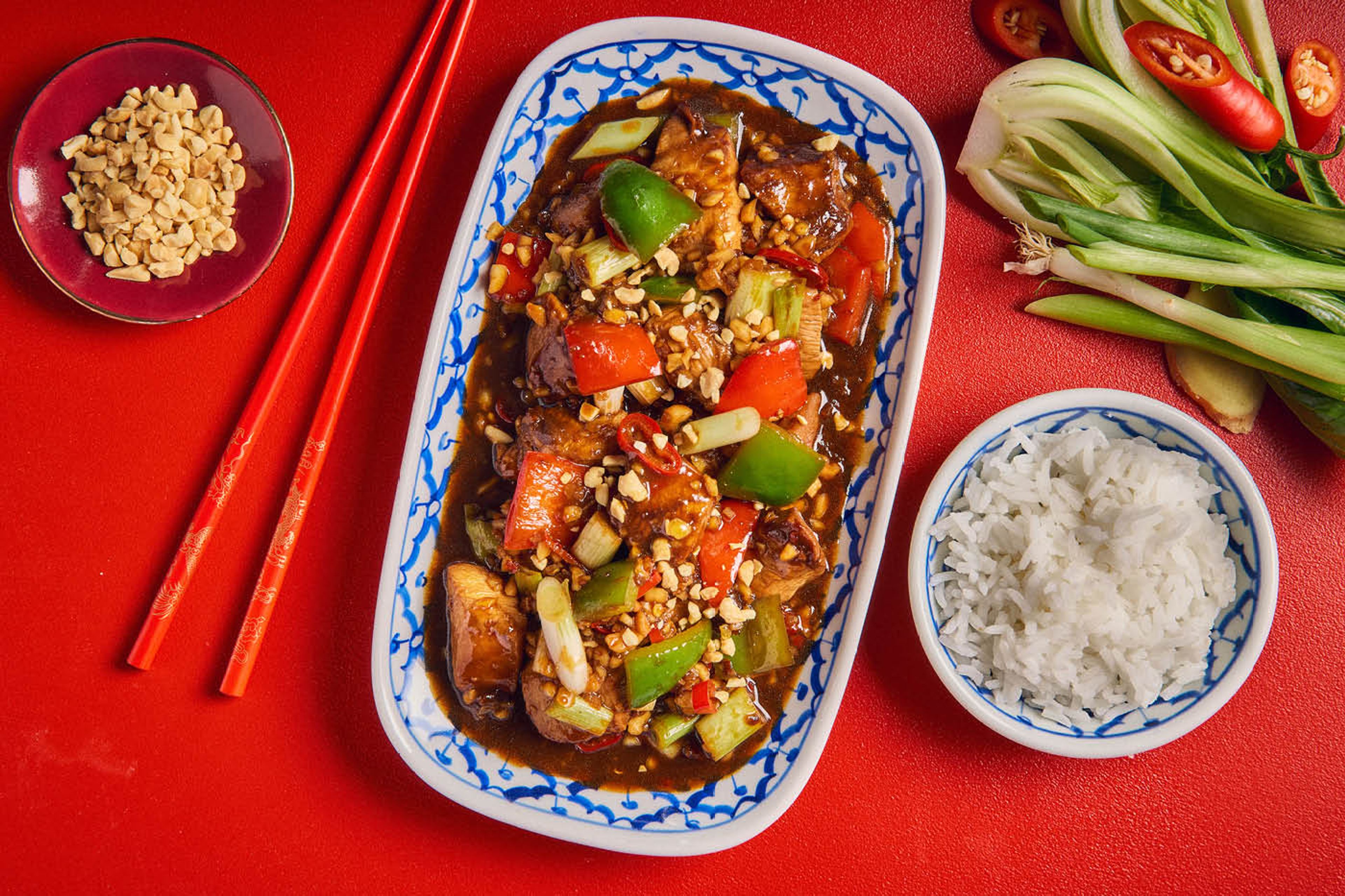 Boller og fat med kung pao chicken og peanøtter mot rød bakgrunn