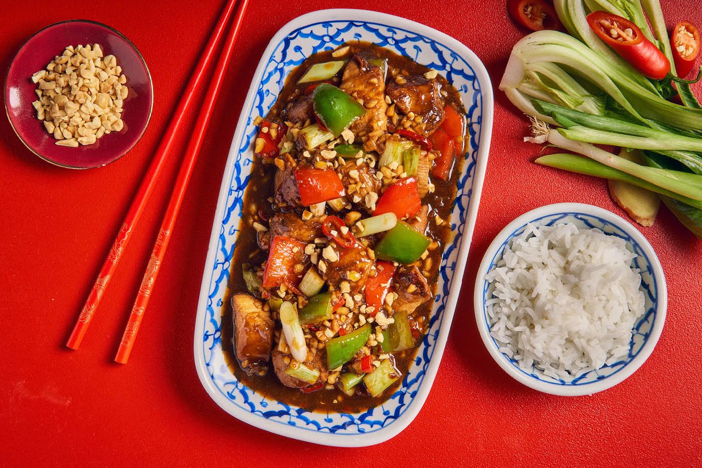 Boller og fat med kung pao chicken og peanøtter mot rød bakgrunn
