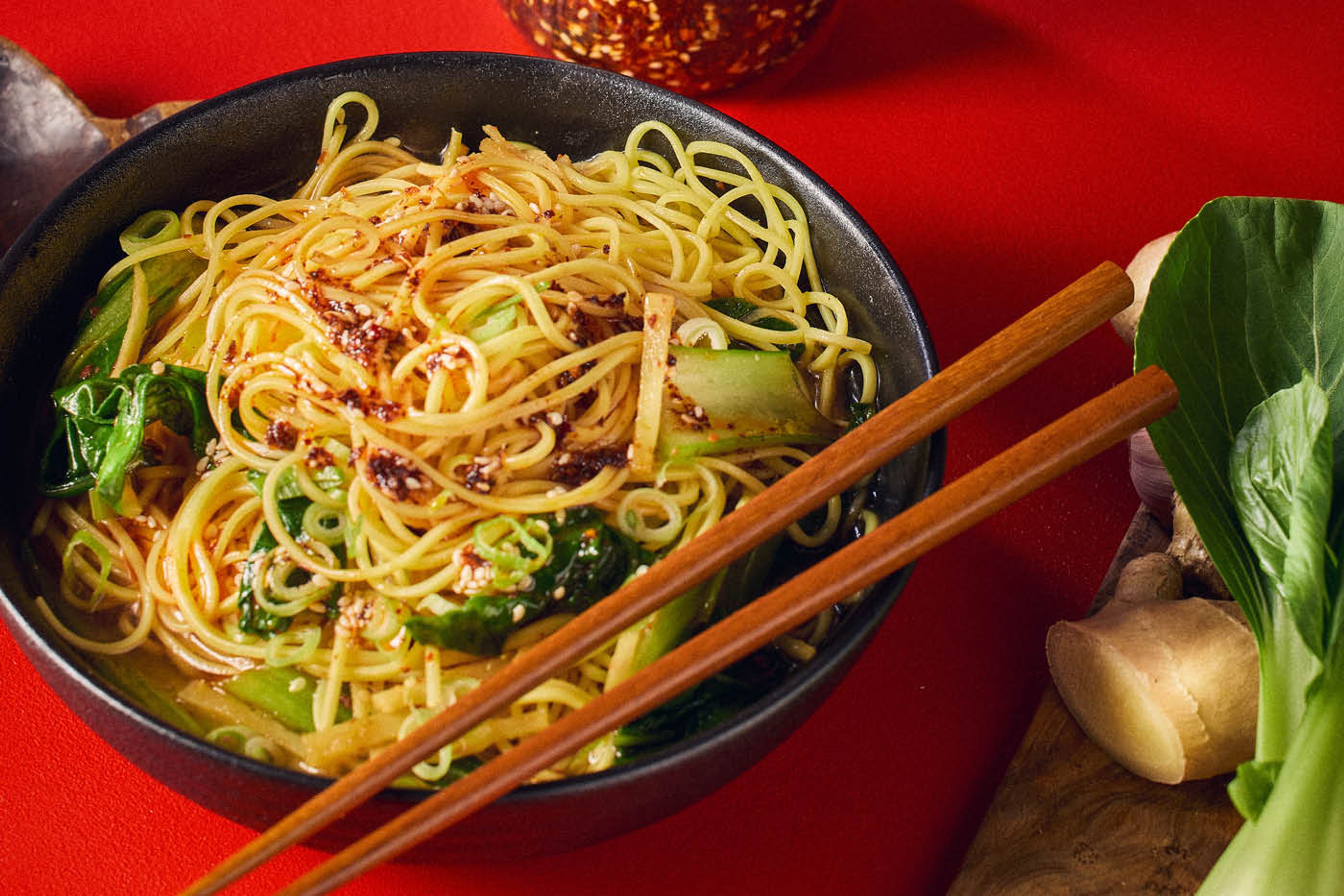 En svart skål fylld med asiatiska nudlar och grönsaker. Ätpinnar på kanten av skålen.