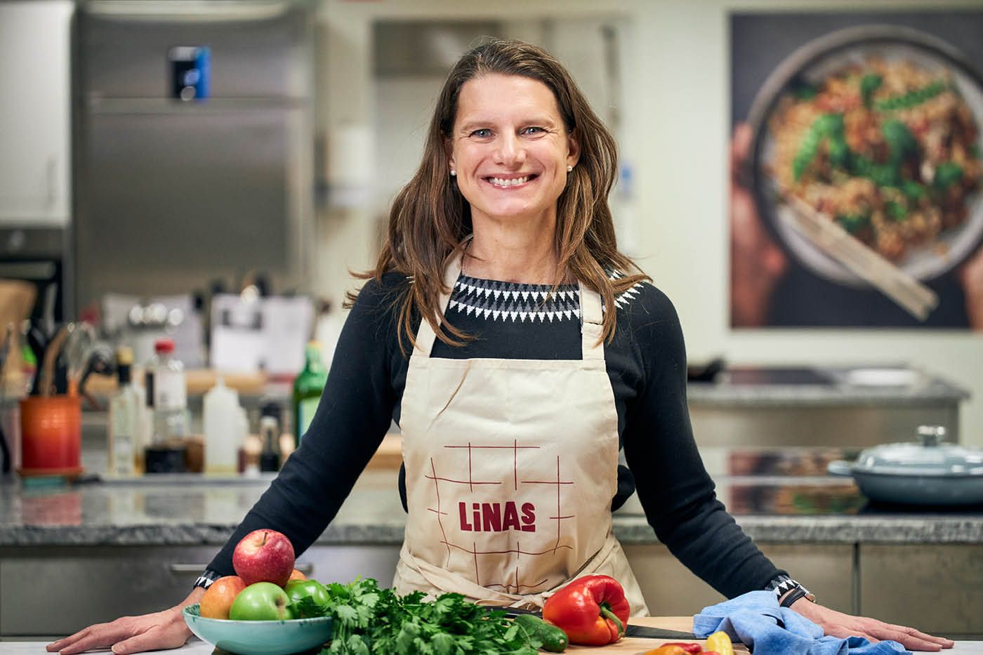 Kvinna i förkläde vid en köksbänk fylld av grönsaker och frukt