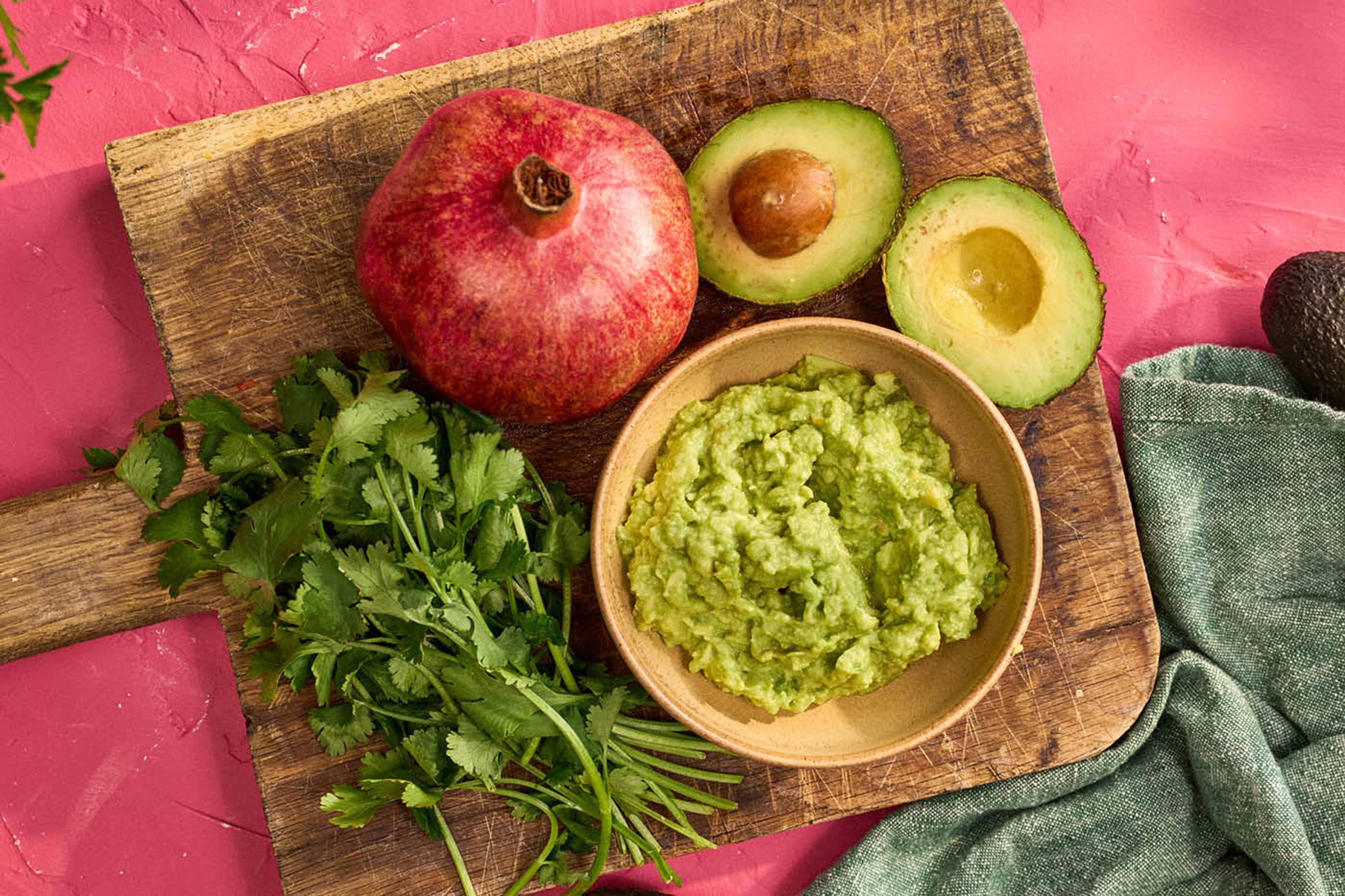 Avokado, koriander och granatäpple på skärbräda mot rosa bakgrund