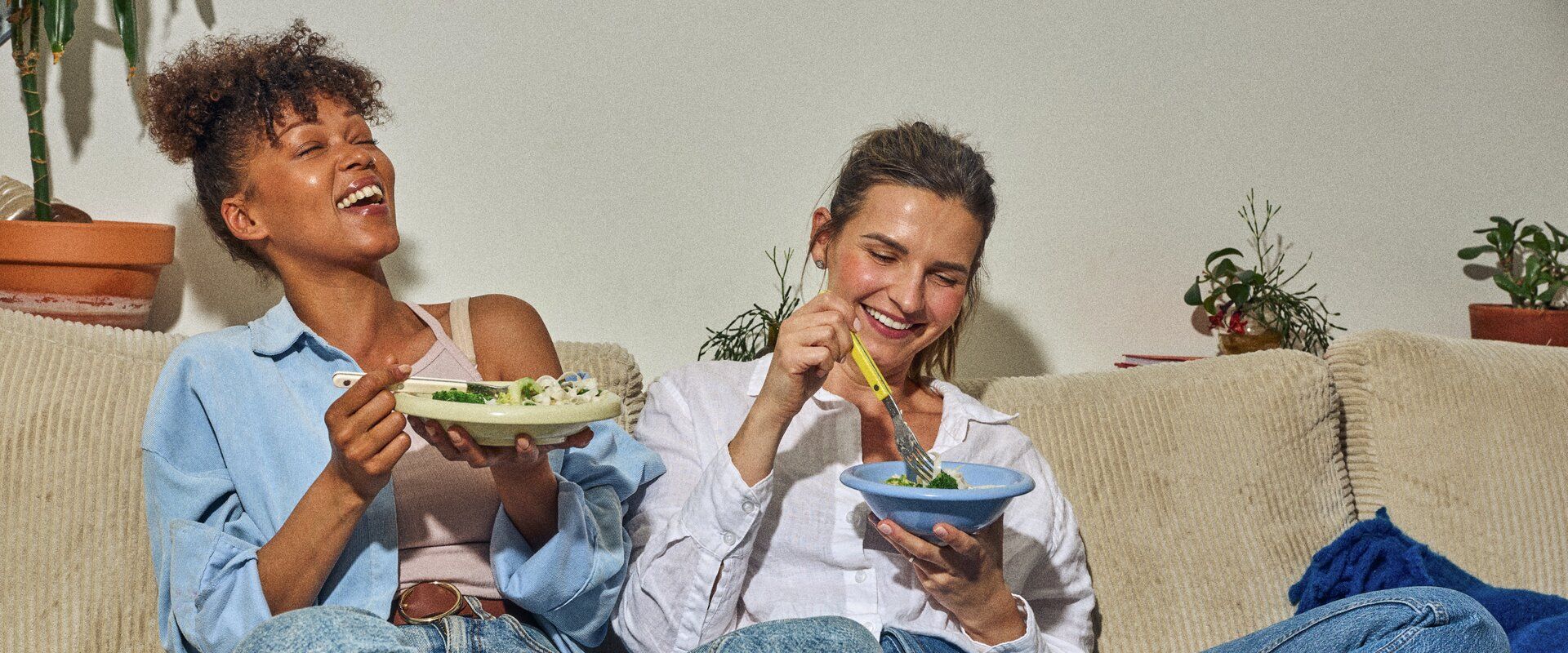 Två kvinnor sitter på en soffa och äter sallad ur skålar.