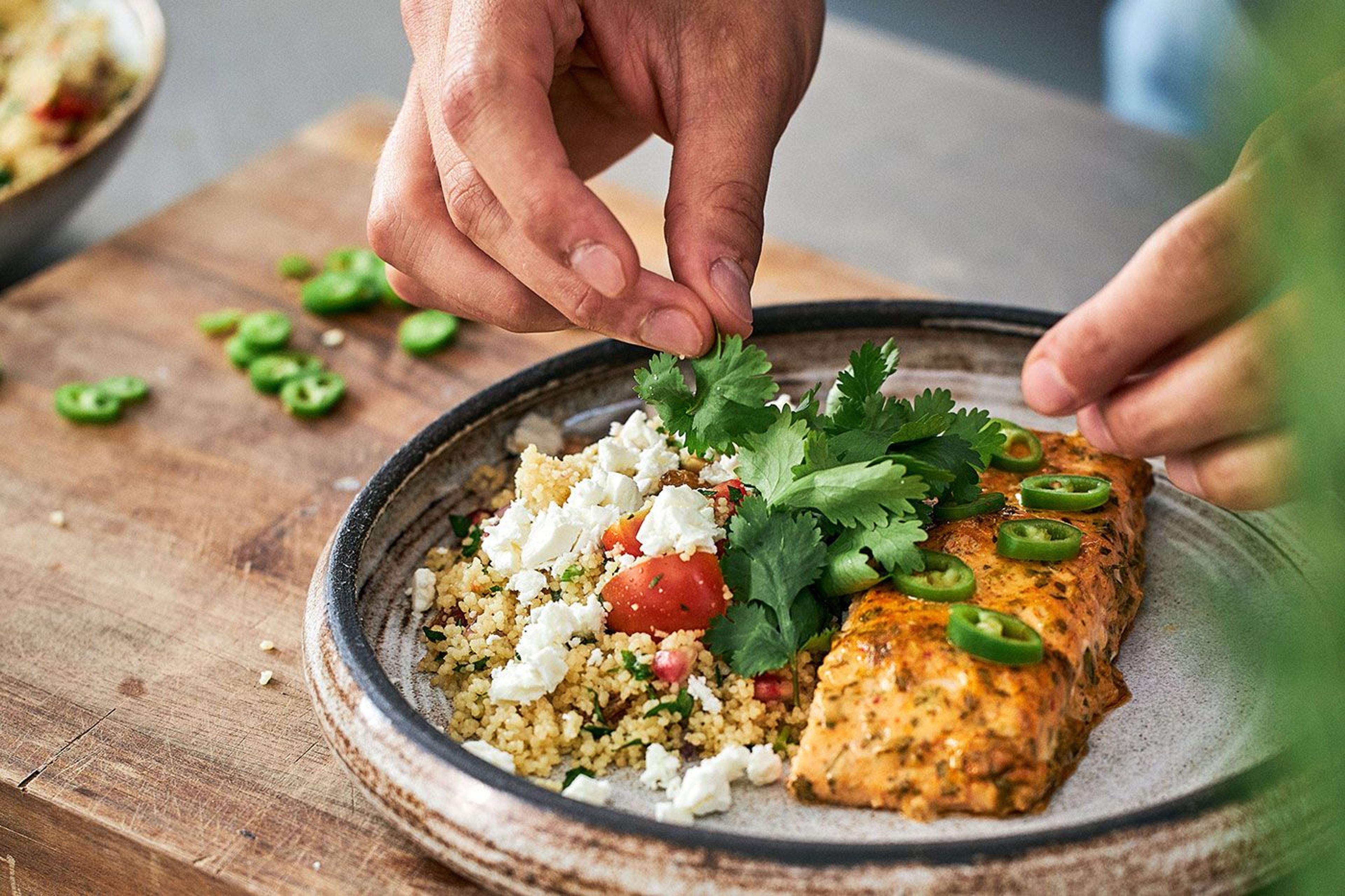 En tallrik med en bit lax, couscous och fetaost. Händer lägger på färska örter