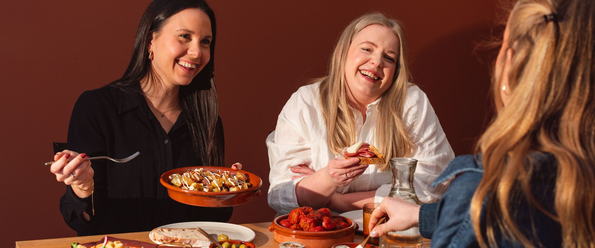 Tre kvinnor sitter runt ett bord, skrattar, lägger upp mat och äter.