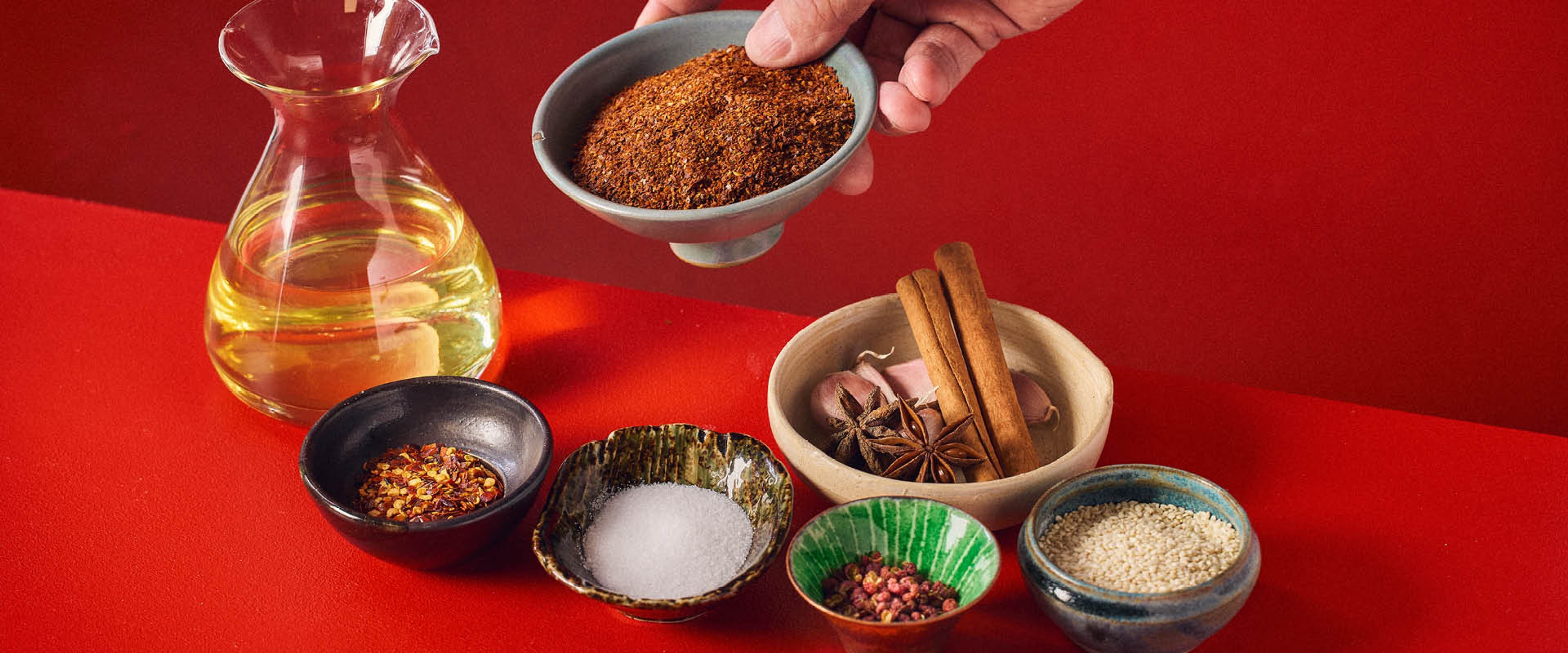 En karaff med olja och många små skålar med ingredienser till chiliolja, som chiliflakes, kanelstänger, stjärnanis, sesamfrön, 