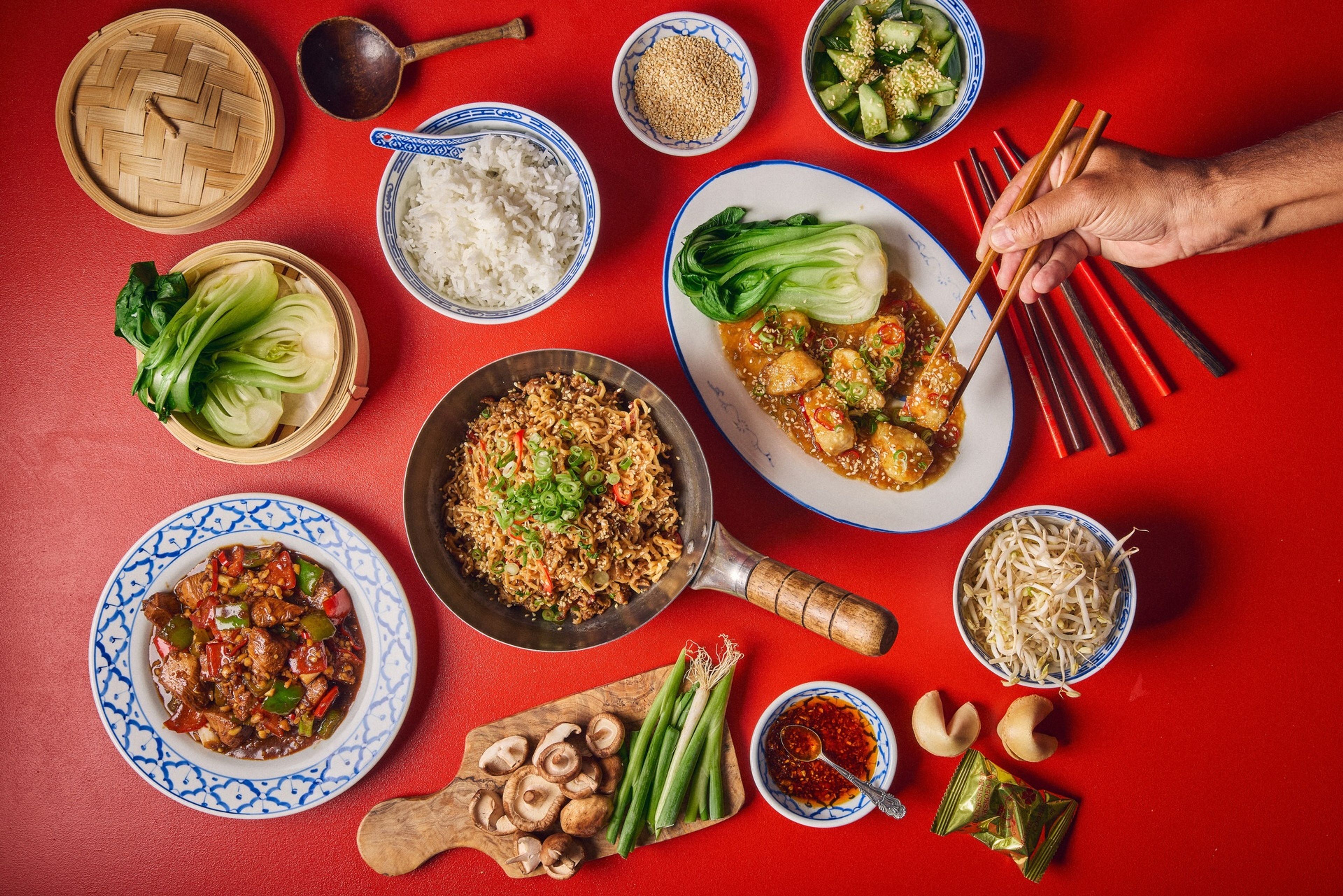 Skålar, skärbrädor och en wokpanna fyllda med råvaror och kinesiska rätter mot röd bakgrund