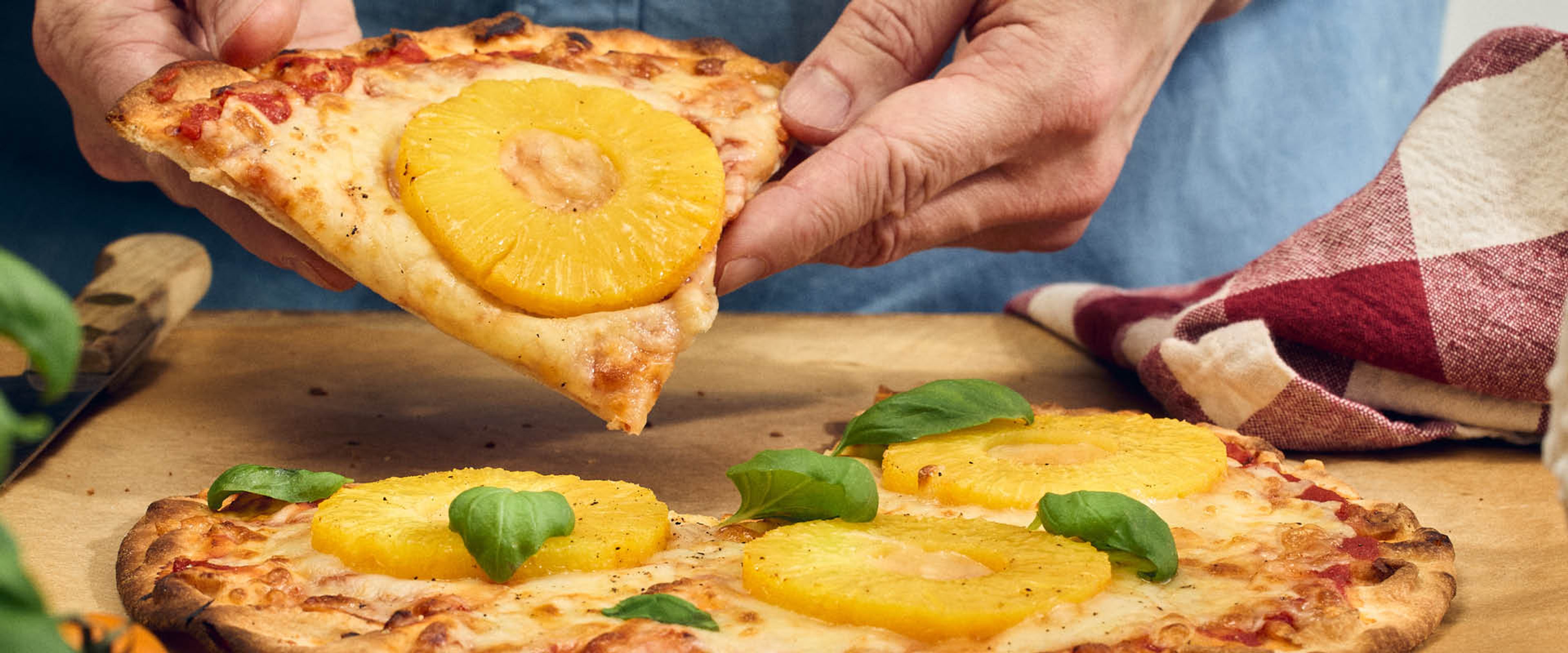 En hånd løfter et pizzastykke toppet med ananas, ost og friske basilikumblade