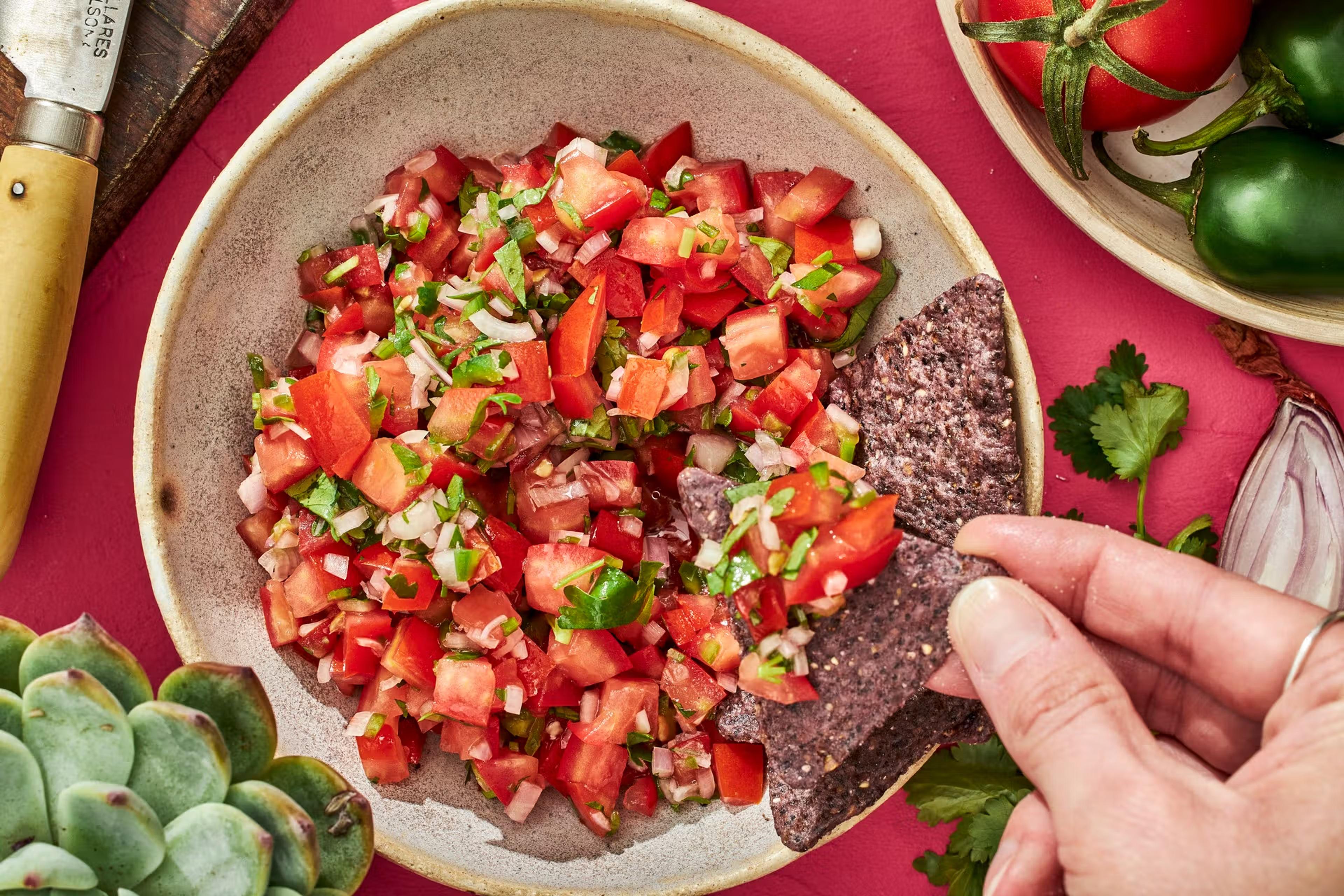 Hånd der holder en tortillachip og dipper i en skål med pico de gallo.