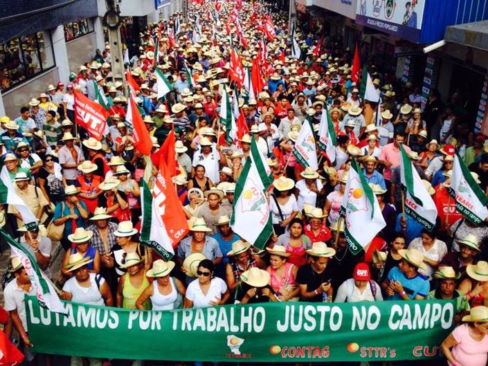 Marcha de trabalhadores e trabalhadoras rurais com bandeiras sindicais, sob a faixa "Lutamos por trabalho justo no campo".