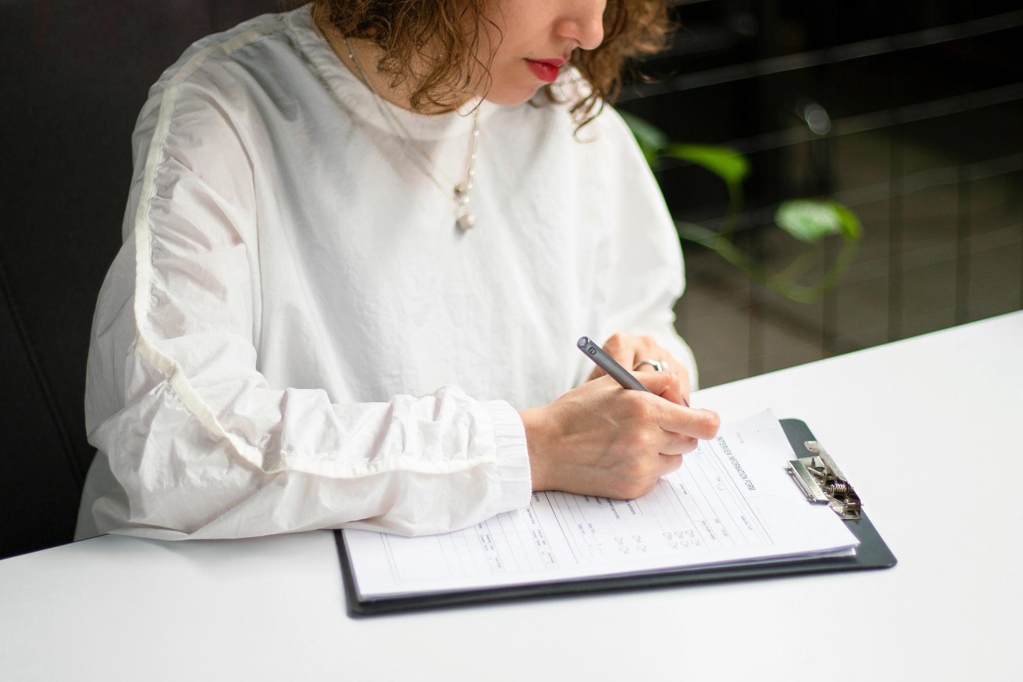 A woman sitting at a table writing onto a piece of paper in a clipboard