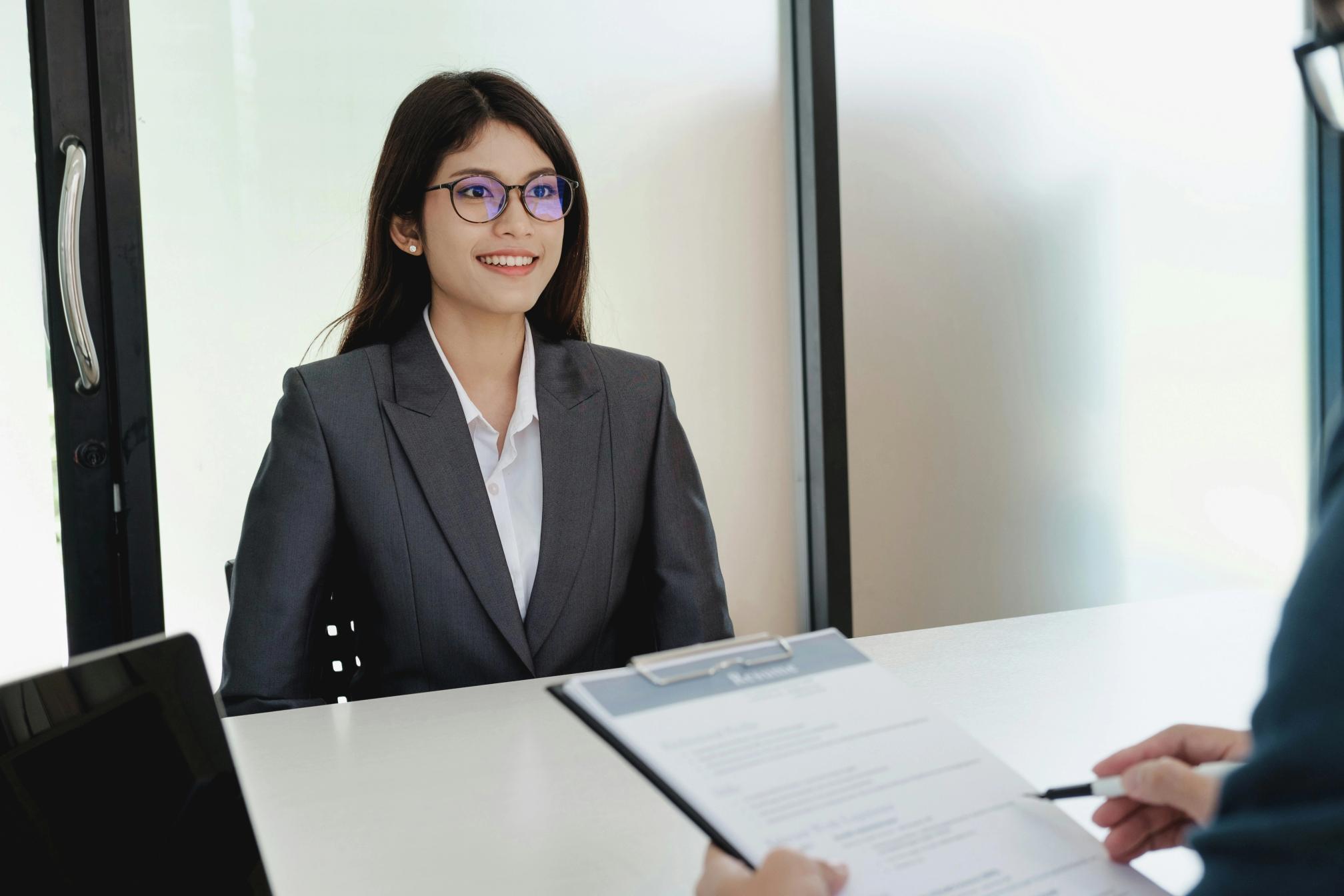 A woman in a suit and glasses being interviewed by a man holding a resume