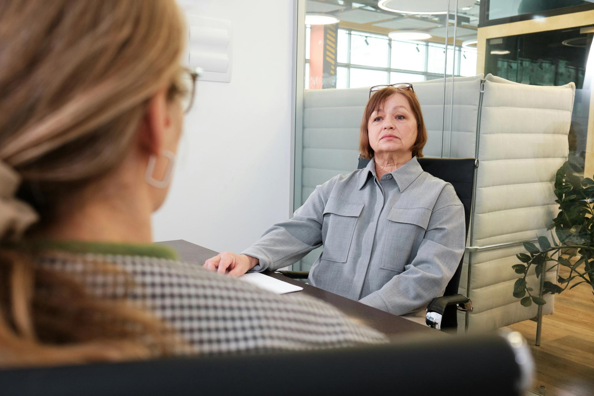 A blonde woman with glasses and earrings being interviewed by another woman in a corporate office.