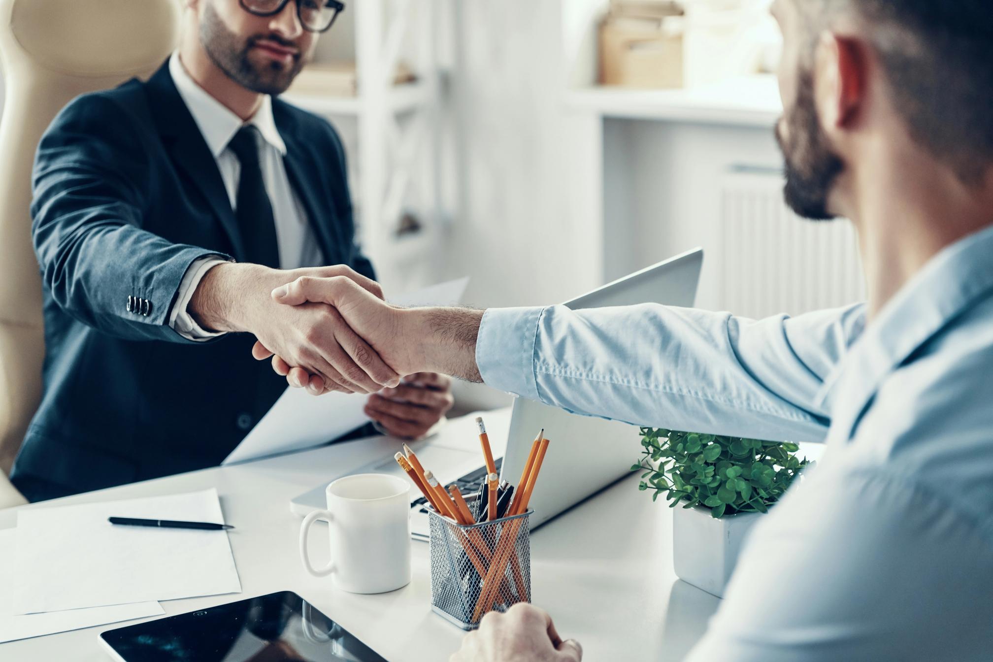 Two formally dressed men in a modern corporate setting shaking hands over a desk