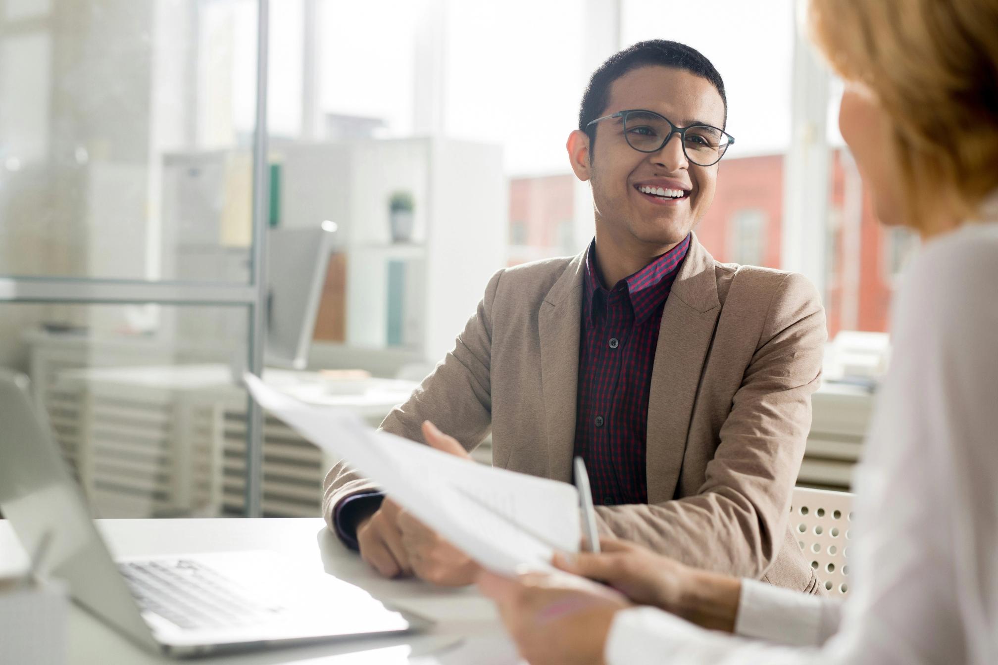A woman interviewing a smiling man with glasses in a suit in a corporate office.