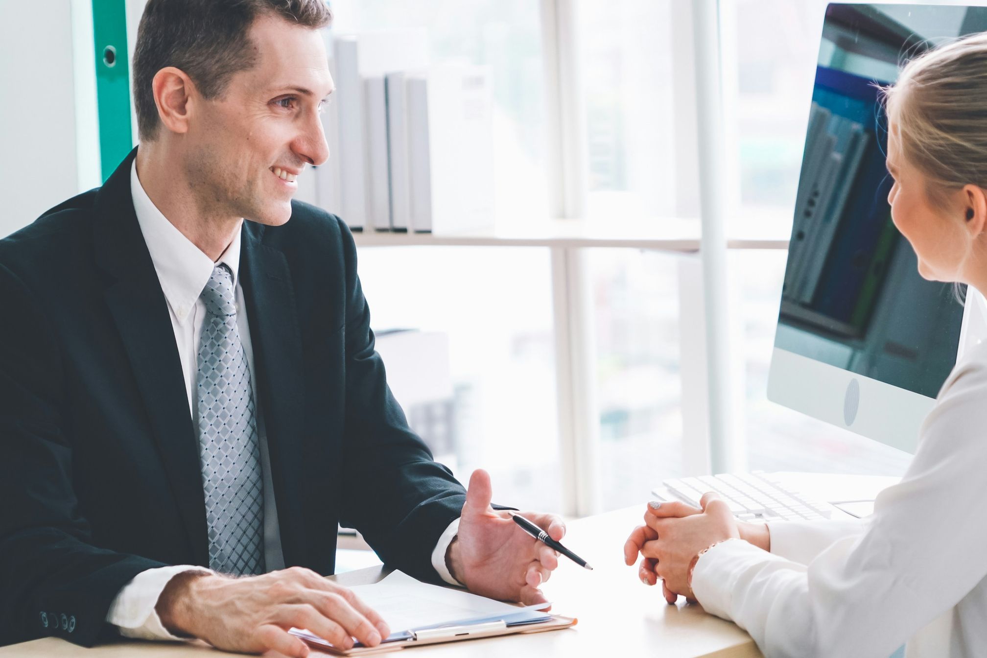 A man in a suit with a pen and paper interviewing a blonde woman in an office setting