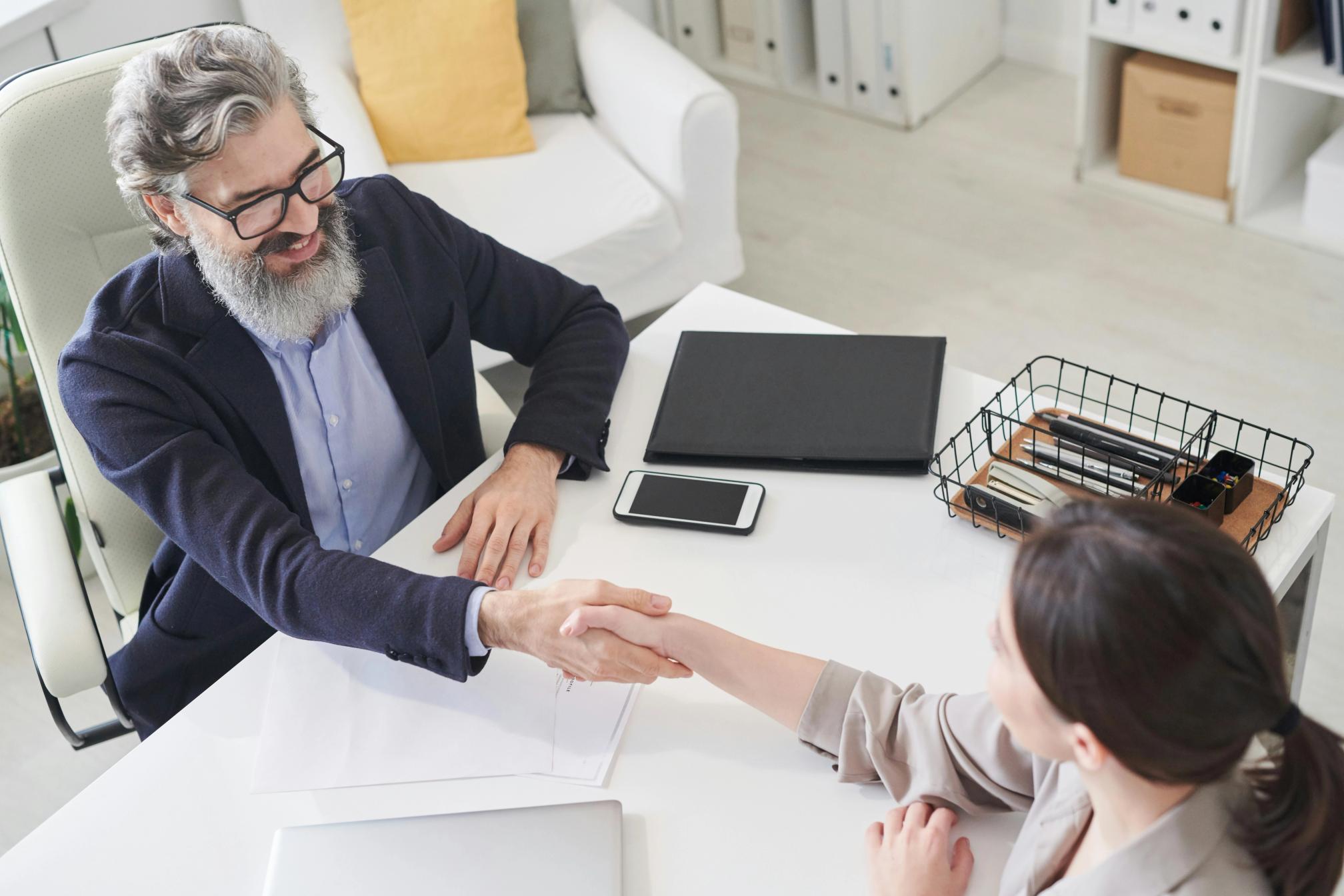 A bearded man with glasses in a suit shaking hands with a woman over a desk in a modern corporate setting