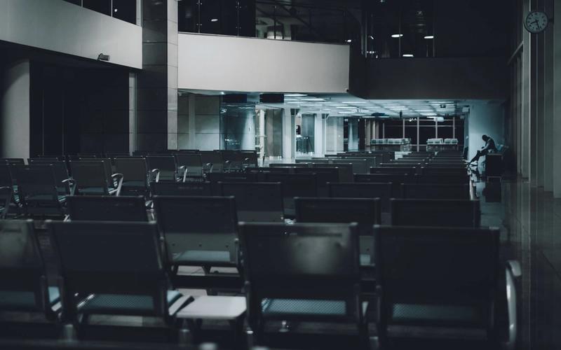 A dimly lit airport waiting area with empty rows and a lone figure in the distance.
