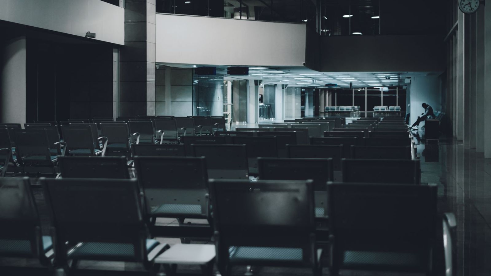 A dimly lit airport waiting area with empty rows and a lone figure in the distance.