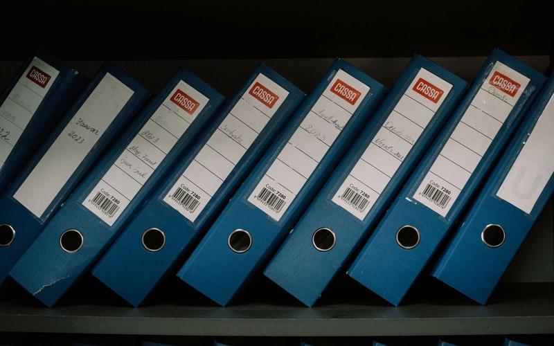 Blue ring binders with handwritten month labels standing in a row on a dark archive shelf.