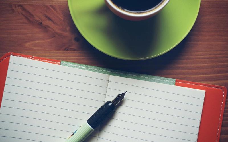 A cup of coffee, a fountain pen, and an open notebook on a wooden surface.