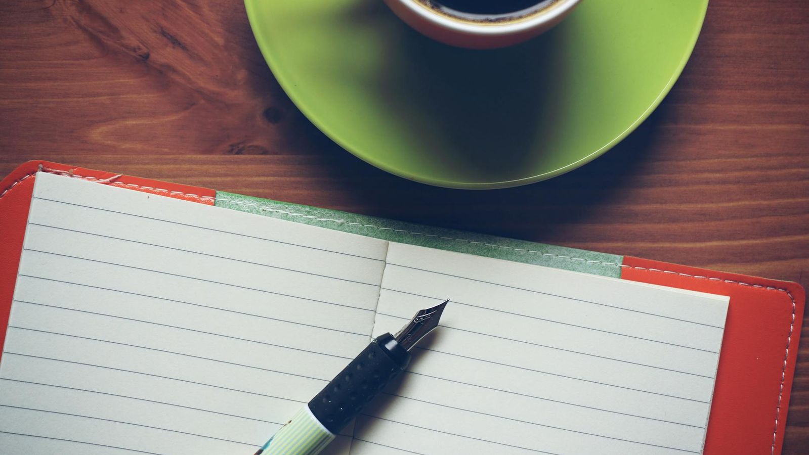En kop kaffe, en fyldepen og en åben notesbog på en træoverflade.