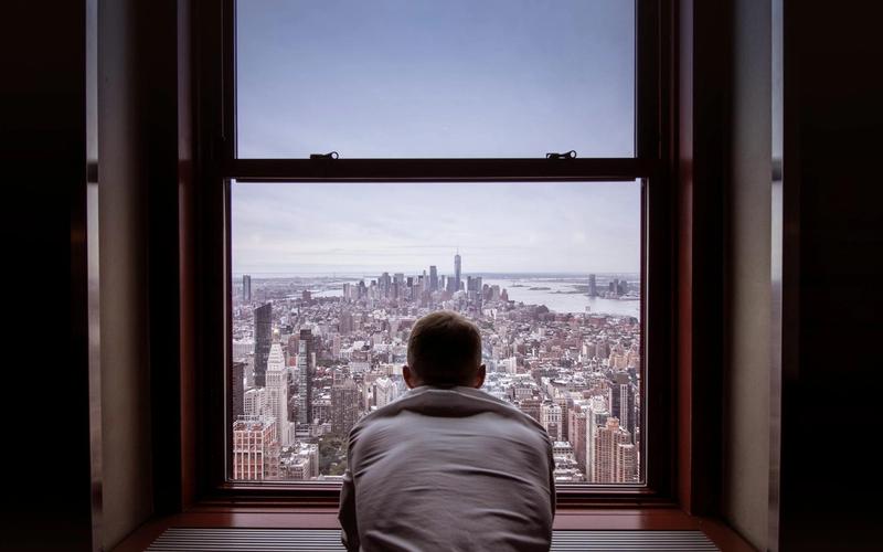 A man stands by a large window overlooking a city skyline in daylight.