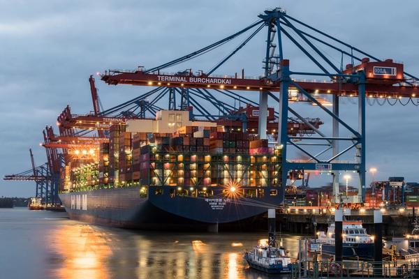 A container ship being loaded at Terminal Burchardkai in Hamburg at dusk, with cranes reaching over the deck.