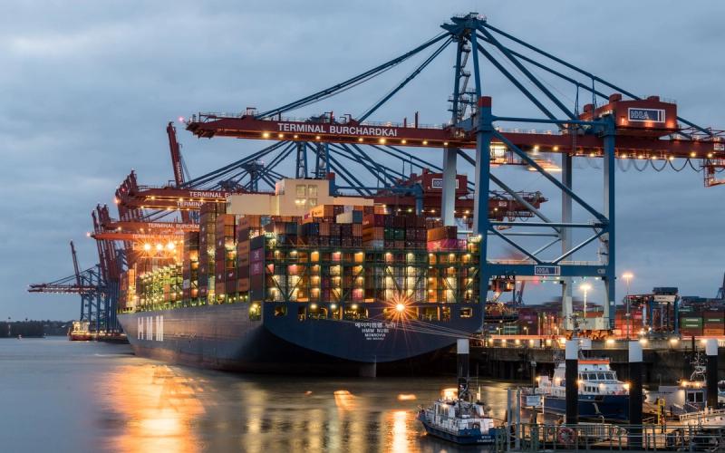 A container ship being loaded at Terminal Burchardkai in Hamburg at dusk, with cranes reaching over the deck.
