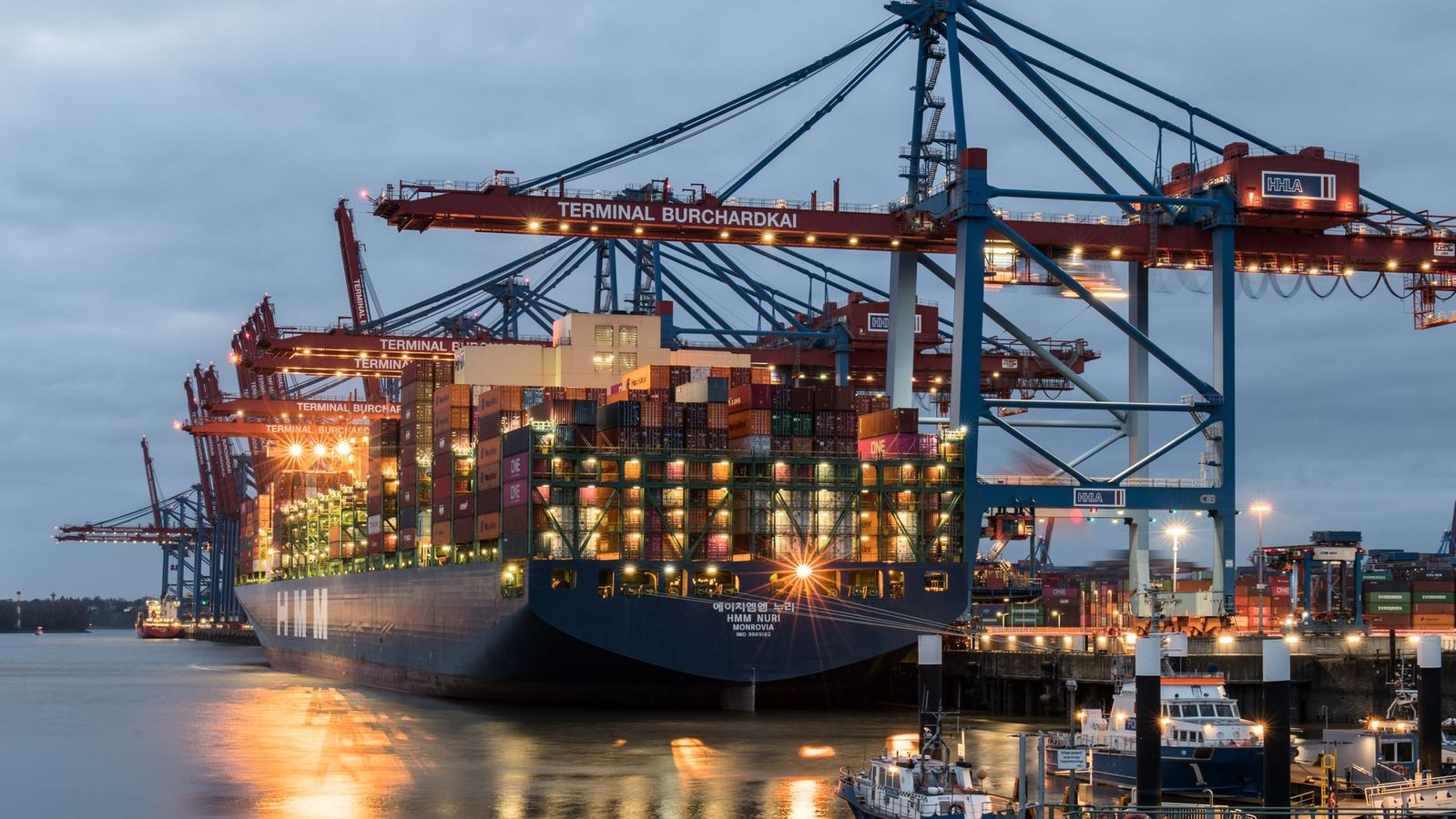 A container ship being loaded at Terminal Burchardkai in Hamburg at dusk, with cranes reaching over the deck.