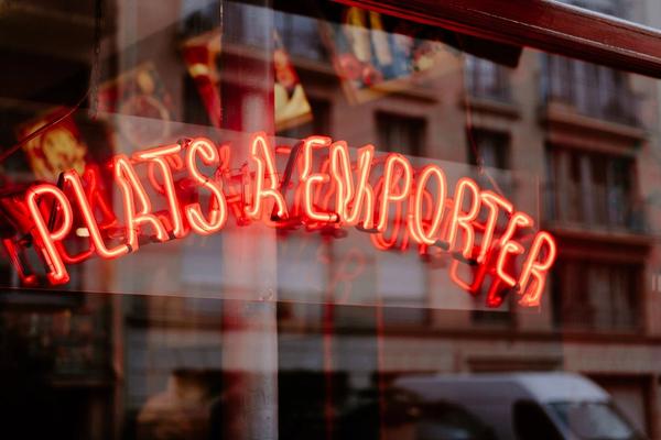 A red neon sign reading Plats a emporter in a shop window, with a Paris facade reflected in the glass.