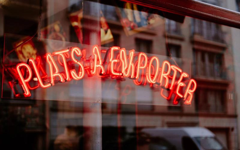 A red neon sign reading Plats a emporter in a shop window, with a Paris facade reflected in the glass.
