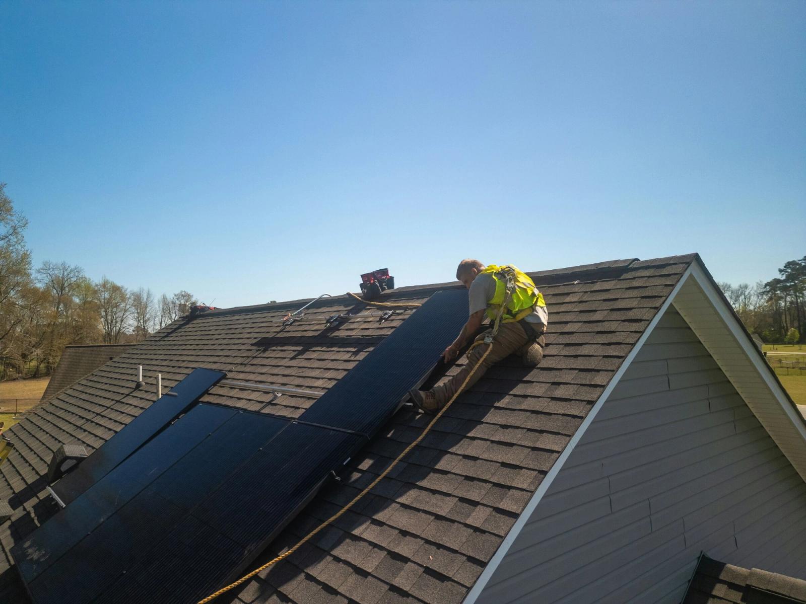 A roofer in a safety vest works on a shingle roof beside a solar array.