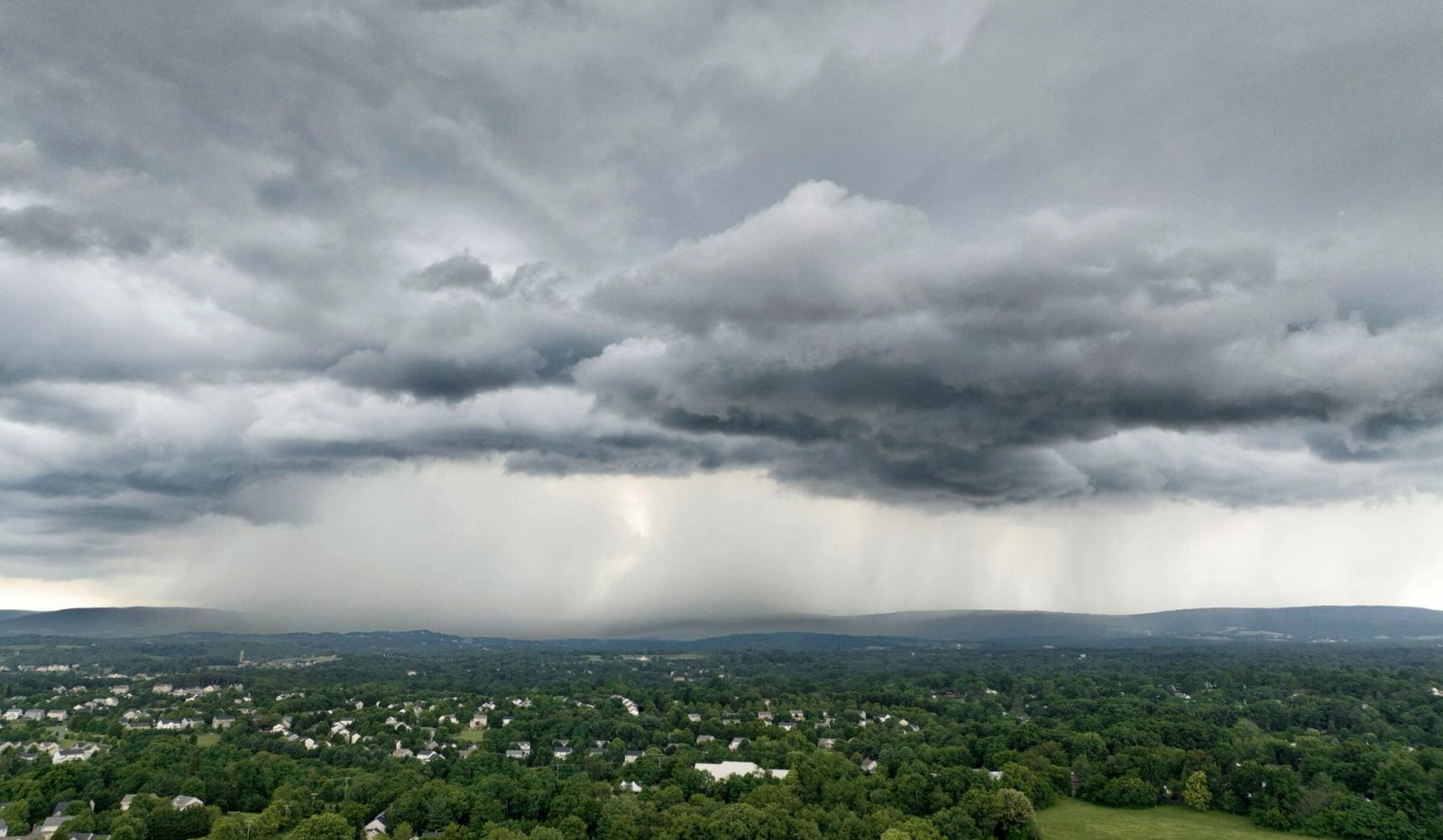 A dark storm cell drops rain over a wooded town, seen from the air.