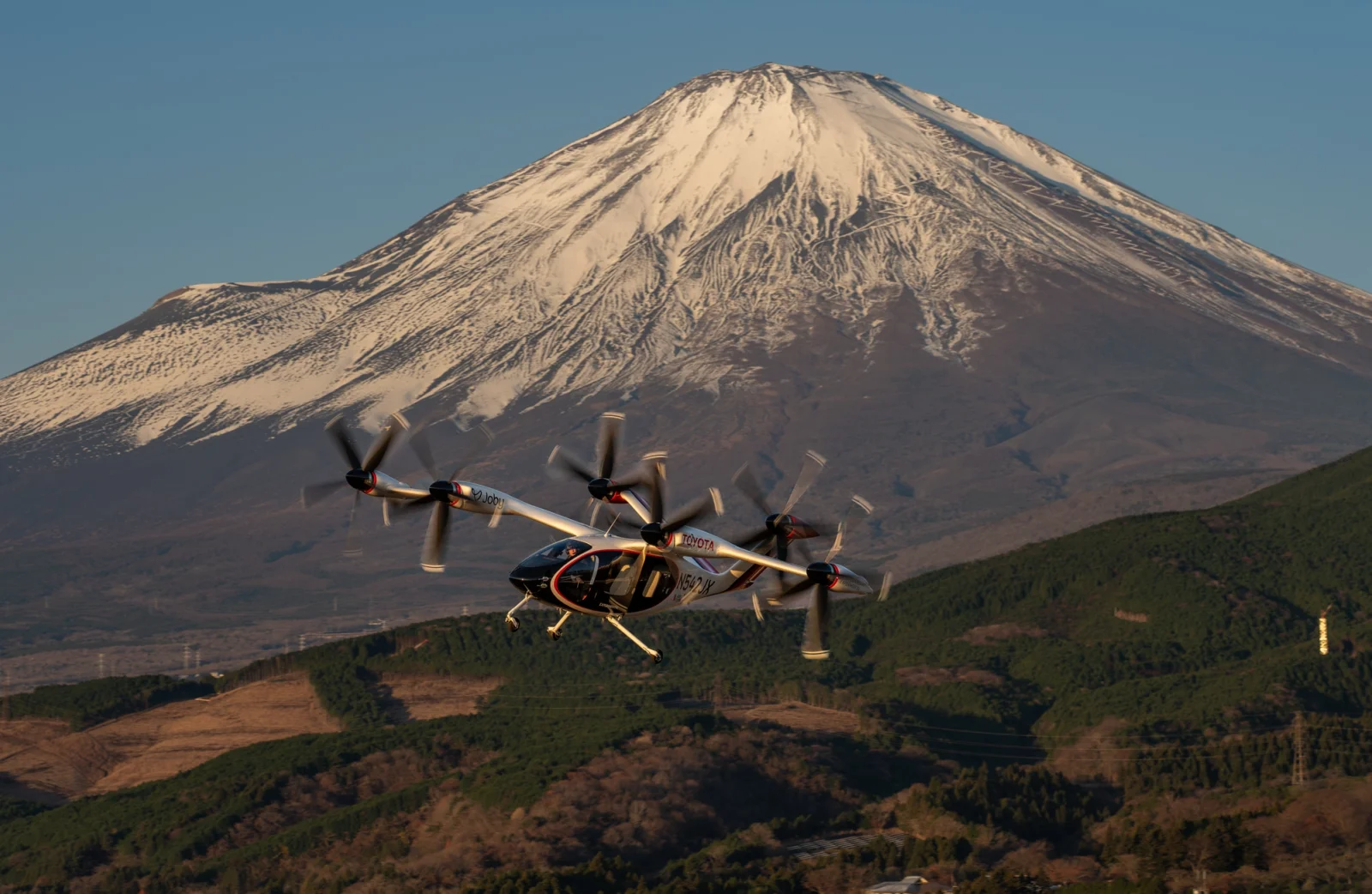The Joby aircraft can be seen flying through the sky with a large mountain in the background.