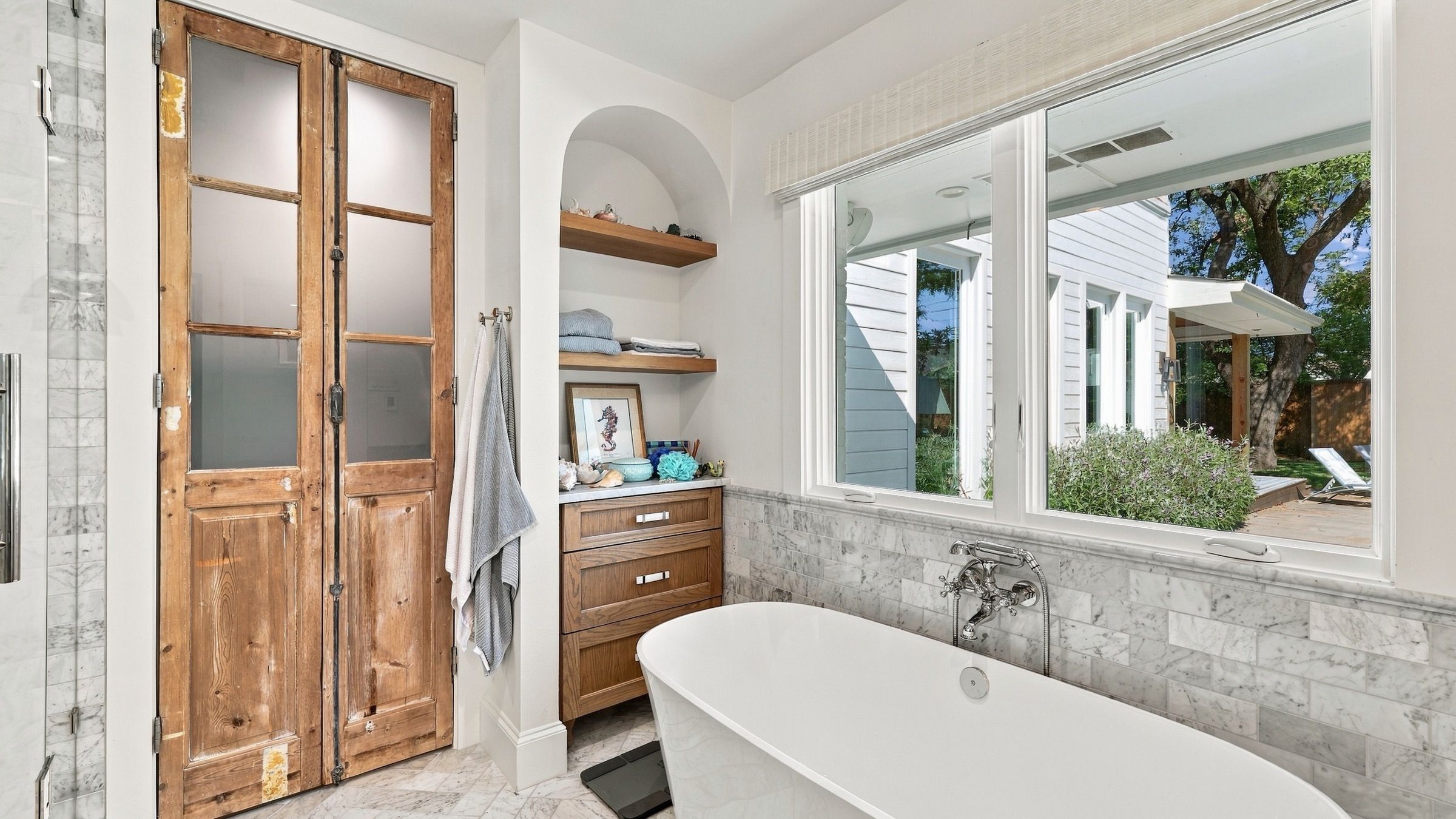 Freestanding tub under a wide window, beside a pair of antique wood doors