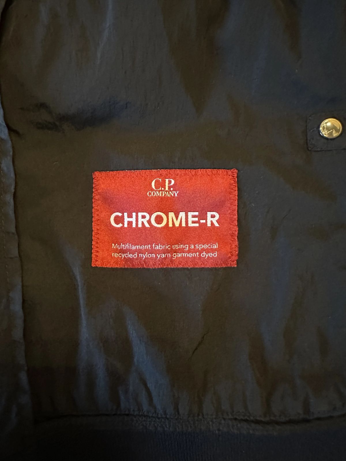 Het rode CHROME-R stoflabel in een C.P. Company jas, met de tekst multifilament fabric using a special recycled nylon yarn garment dyed