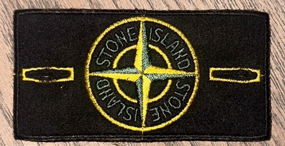 Close-up of a genuine Stone Island compass badge, embroidered in yellow and green on black felt