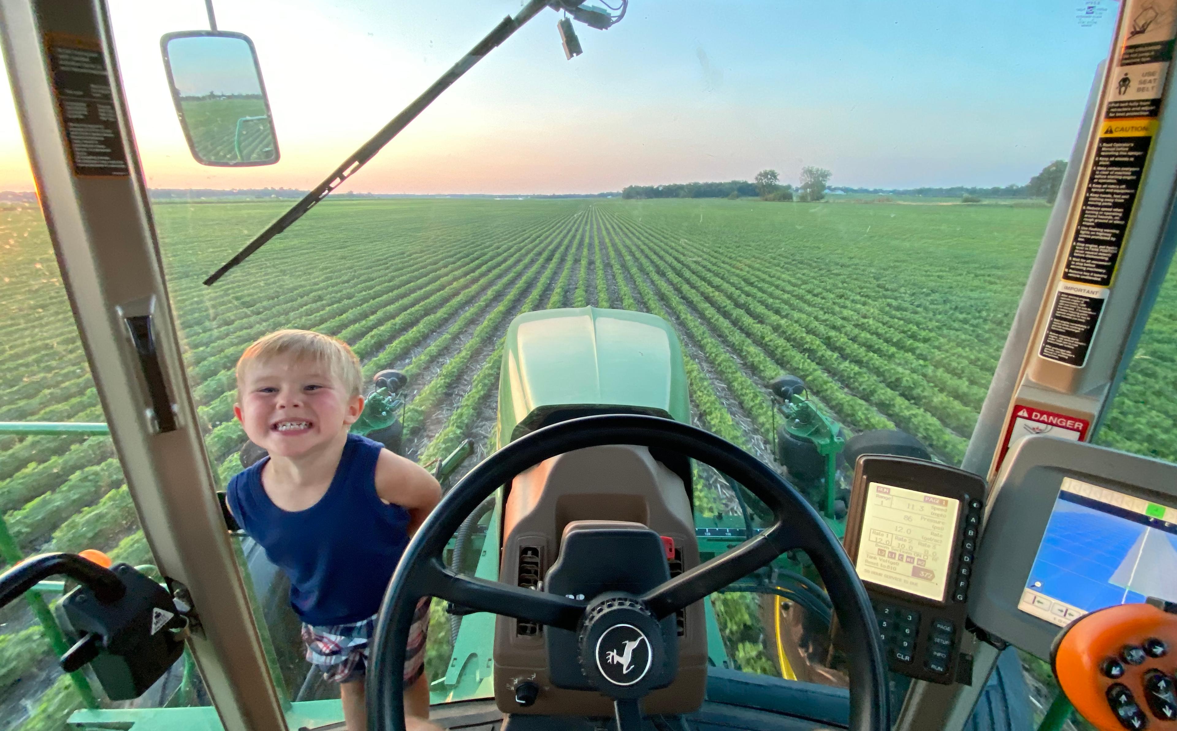 Clark's son grins at the camera from inside the cab of a John Deere tractor. The view from behind the steering wheel looks out over vast, neatly rowed green crop fields — likely soybeans — stretching to the horizon under a pastel sunset sky. GPS navigation screens and safety warning stickers are visible on the cab's interior panels. The tractor's hood and side mirrors are reflected in the wide windshield view.