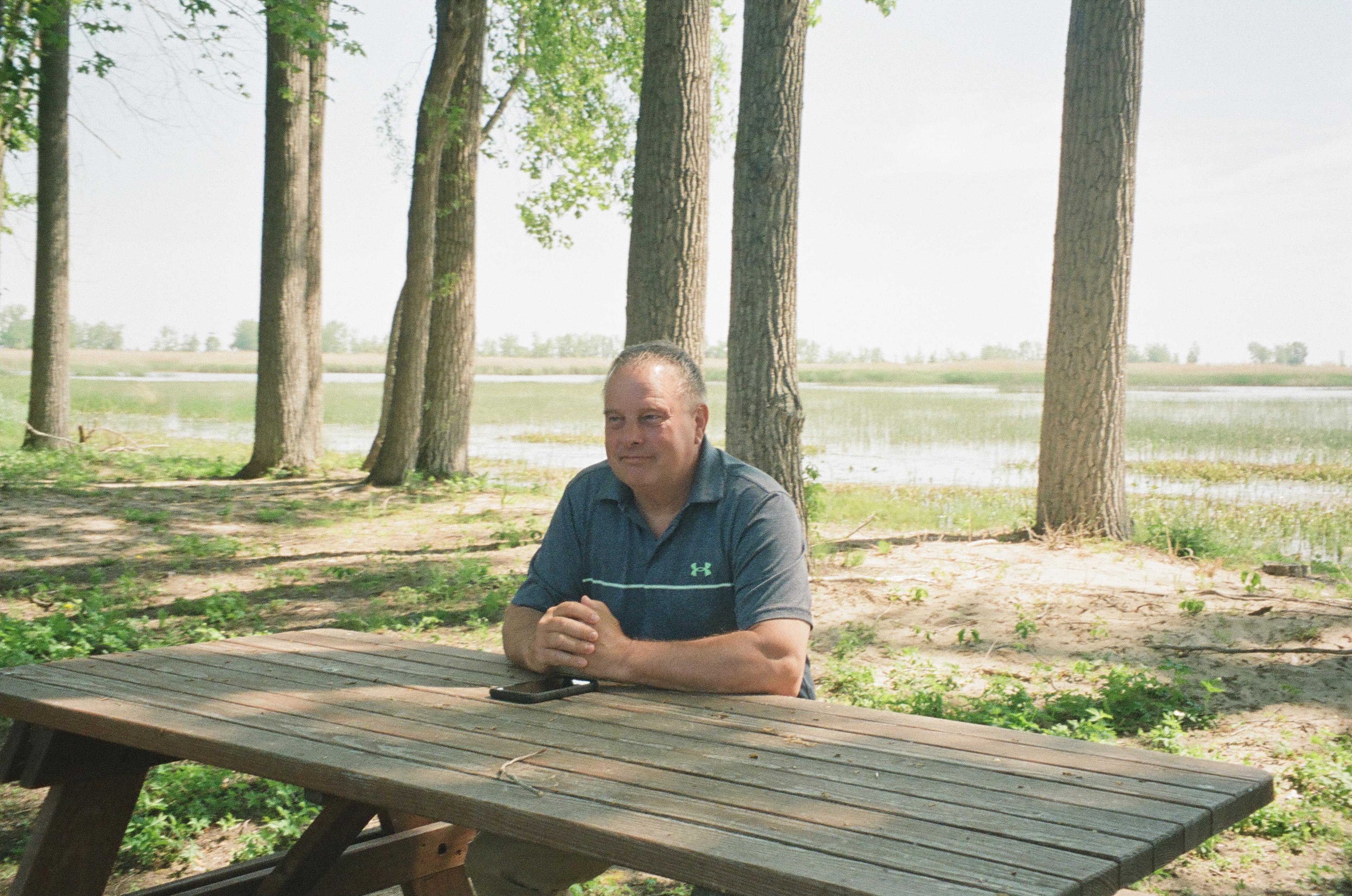 Jeff sits at a wooden lunch table, in the woods near his property.