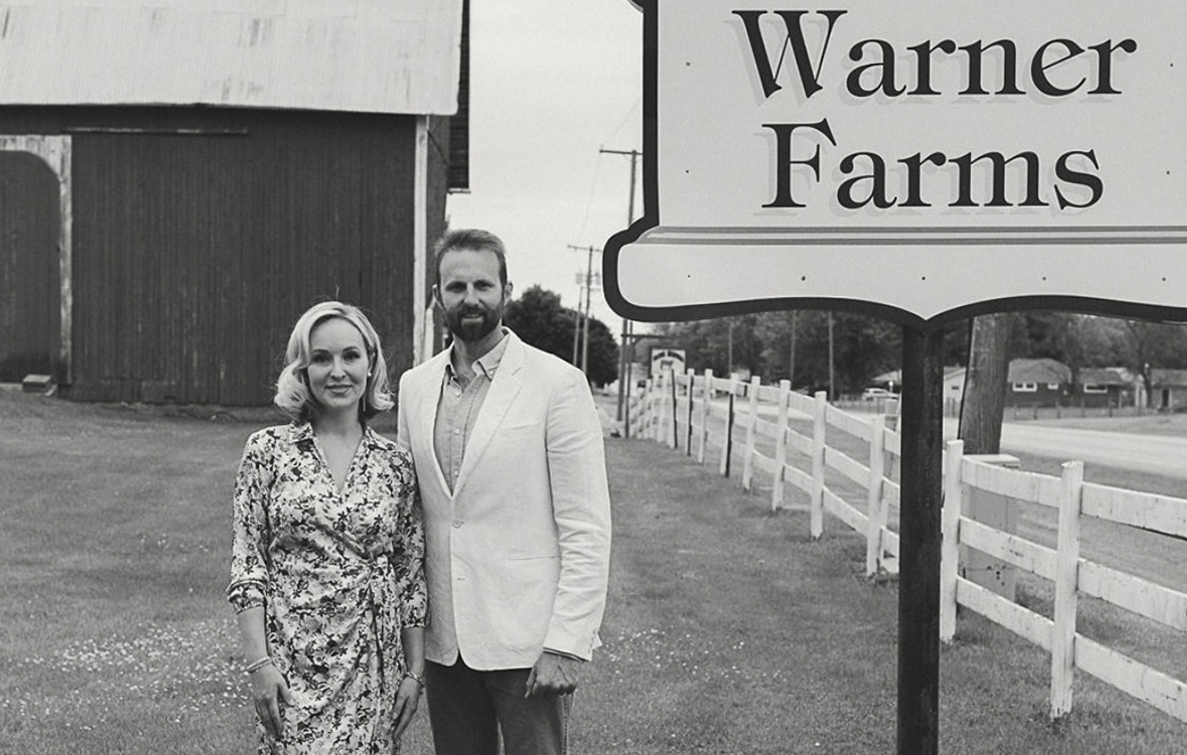 A vintage black-and-white photograph of the Warner family standing beneath a large roadside sign reading "Warner Farms." The woman on the left wears a floral-print shift dress and has short, styled hair. The man on the right wears a light-colored suit jacket. Behind them is a farm barn with silo structures and a white fence, with trees visible in the background.