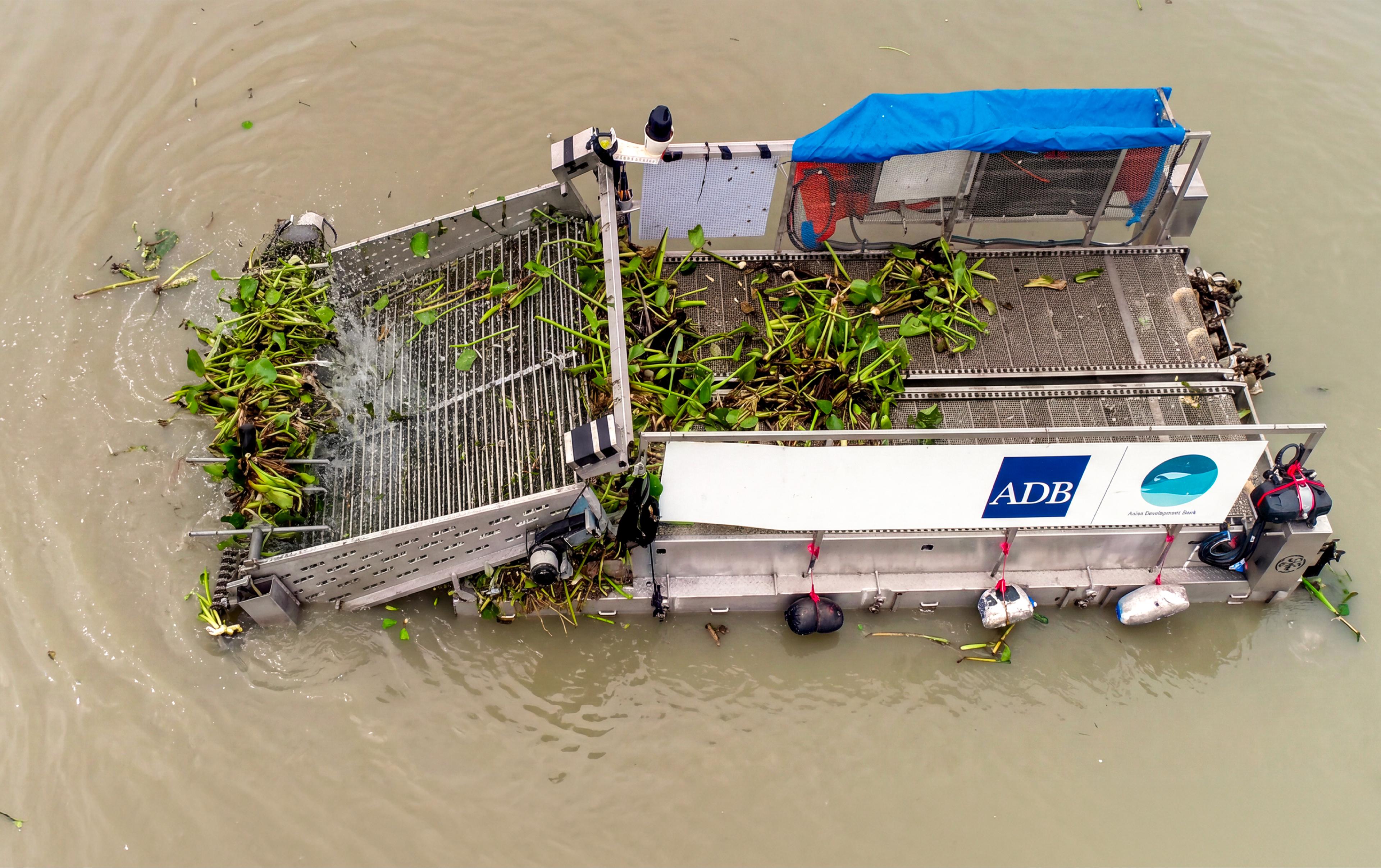 Overhead view of a flat-decked electric harvesting boat on muddy brown water, its conveyor ramp lifting a mat of green water hyacinth onto the deck.