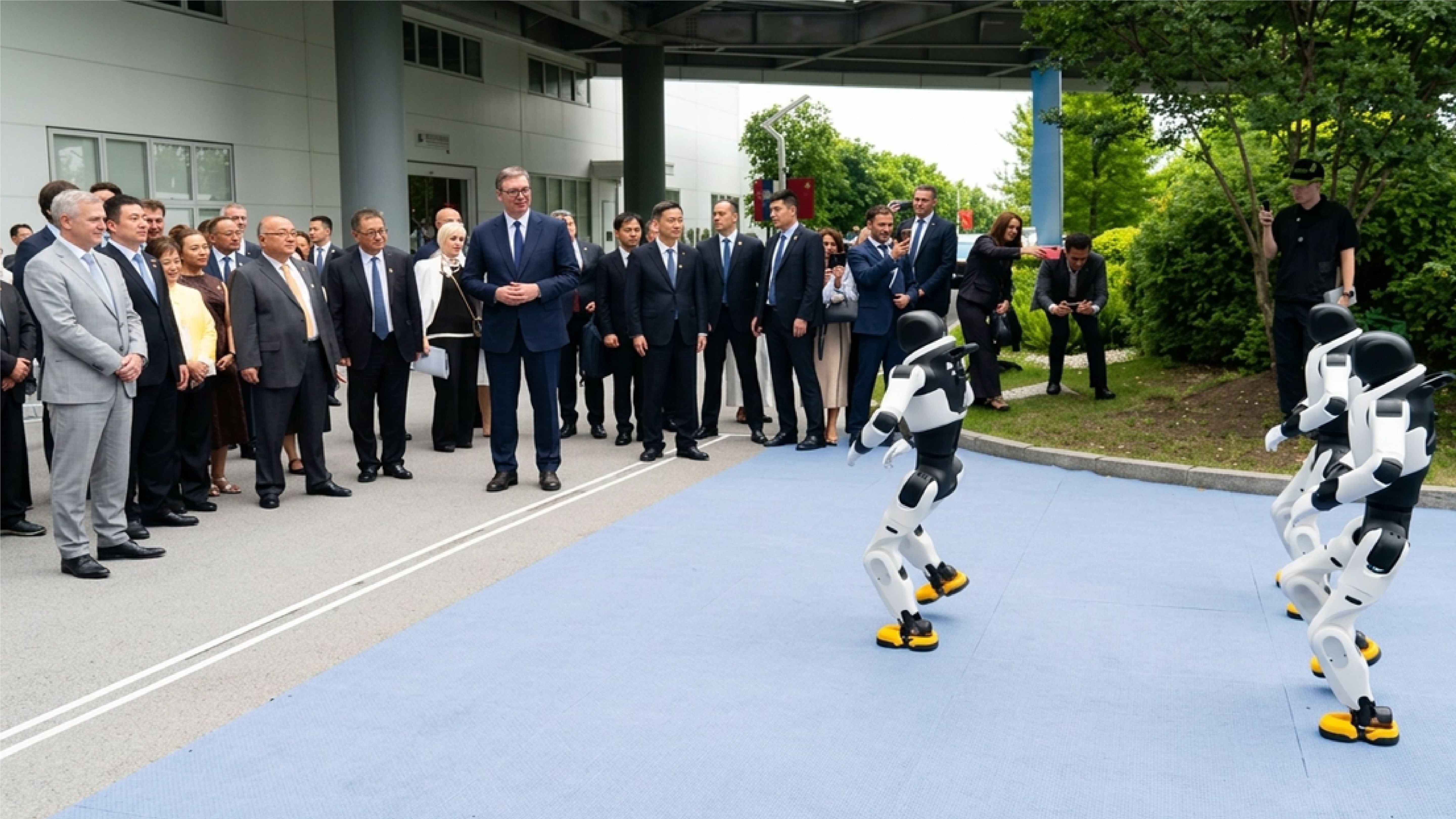 A group of officials in suits watches a demonstration of humanoid robots on an outdoor blue mat. Two white-and-black humanoid robots with yellow feet stand facing the crowd, appearing to perform or move, while onlookers, including several people taking photos with phones, observe from the sidelines near a modern building