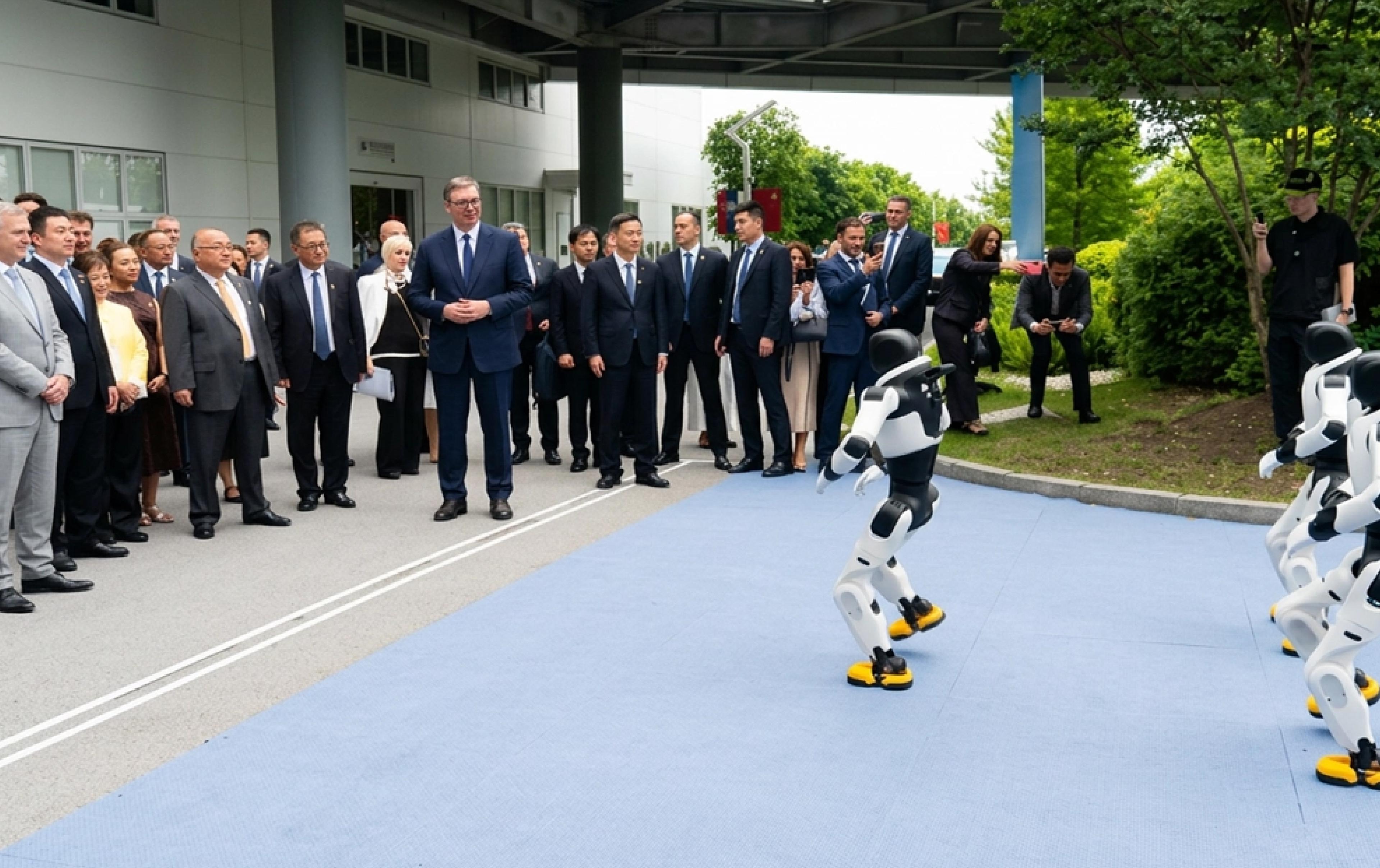 A group of officials in suits watches a demonstration of humanoid robots on an outdoor blue mat. Two white-and-black humanoid robots with yellow feet stand facing the crowd, appearing to perform or move, while onlookers, including several people taking photos with phones, observe from the sidelines near a modern building