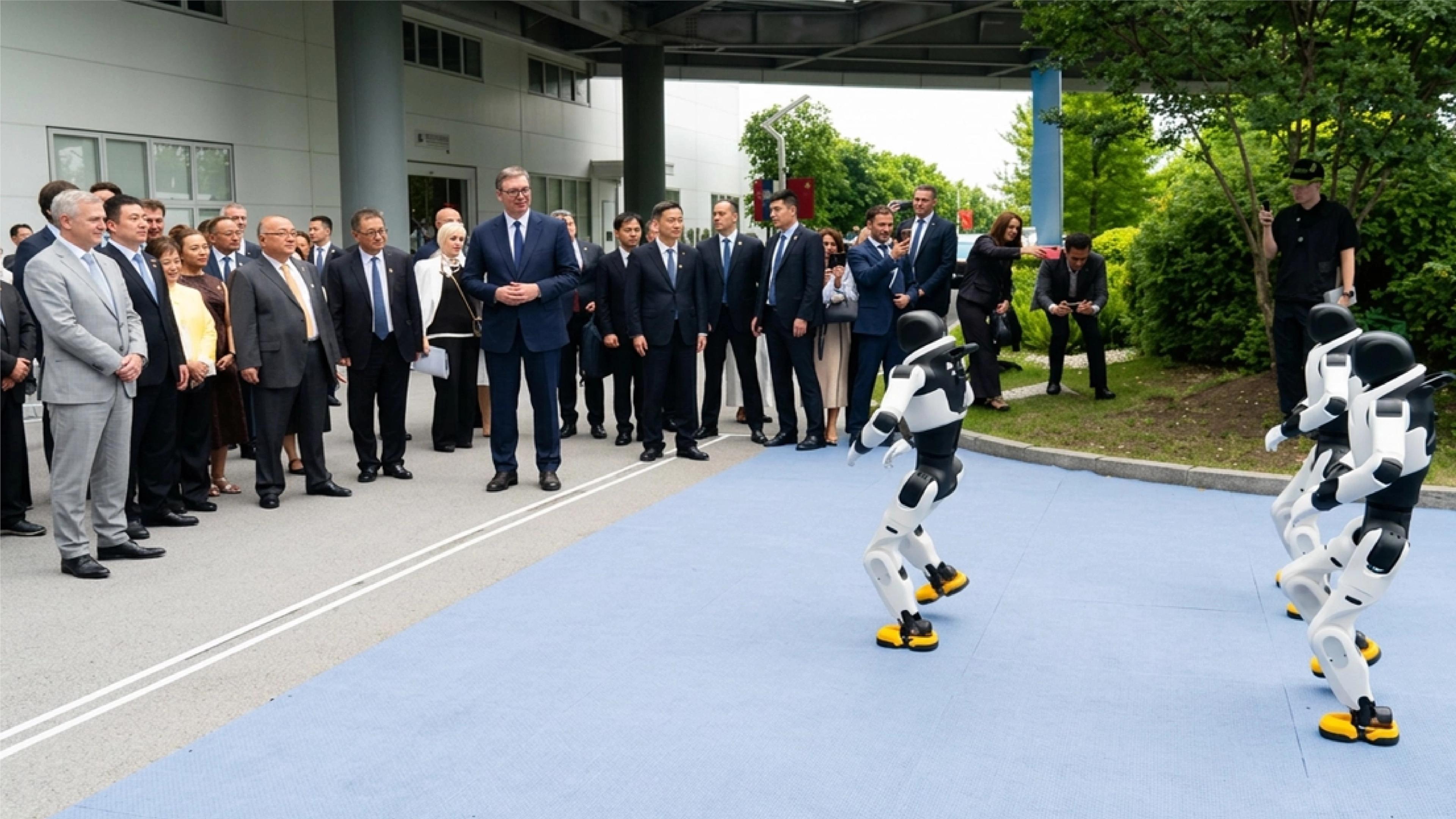 A group of officials in suits watches a demonstration of humanoid robots on an outdoor blue mat. Two white-and-black humanoid robots with yellow feet stand facing the crowd, appearing to perform or move, while onlookers, including several people taking photos with phones, observe from the sidelines near a modern building