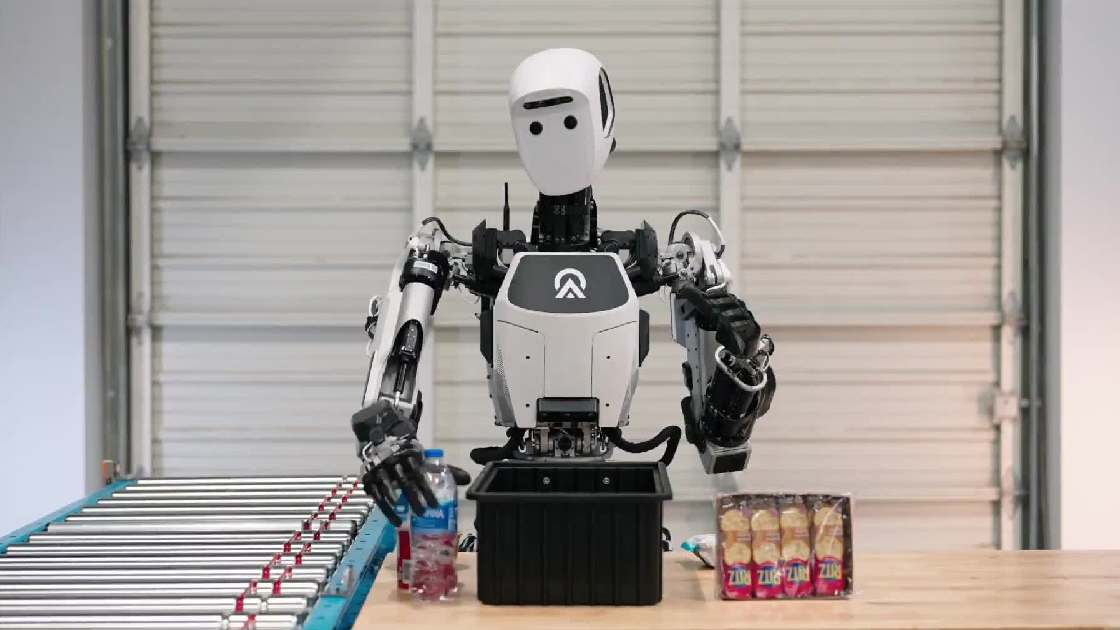 A humanoid robot works at a warehouse-style packing station, moving items between a table, bin and conveyor belt.