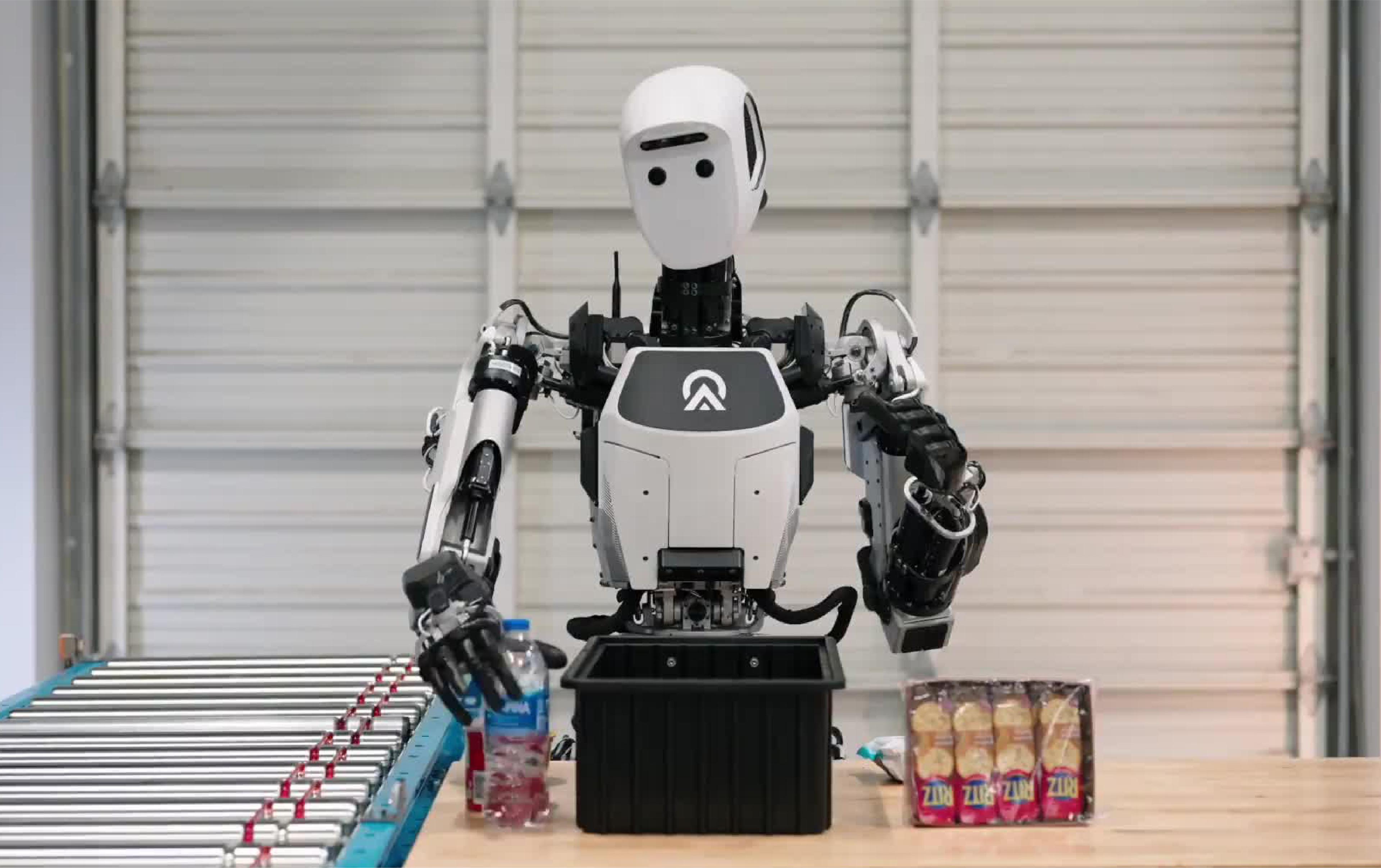A humanoid robot works at a warehouse-style packing station, moving items between a table, bin and conveyor belt.