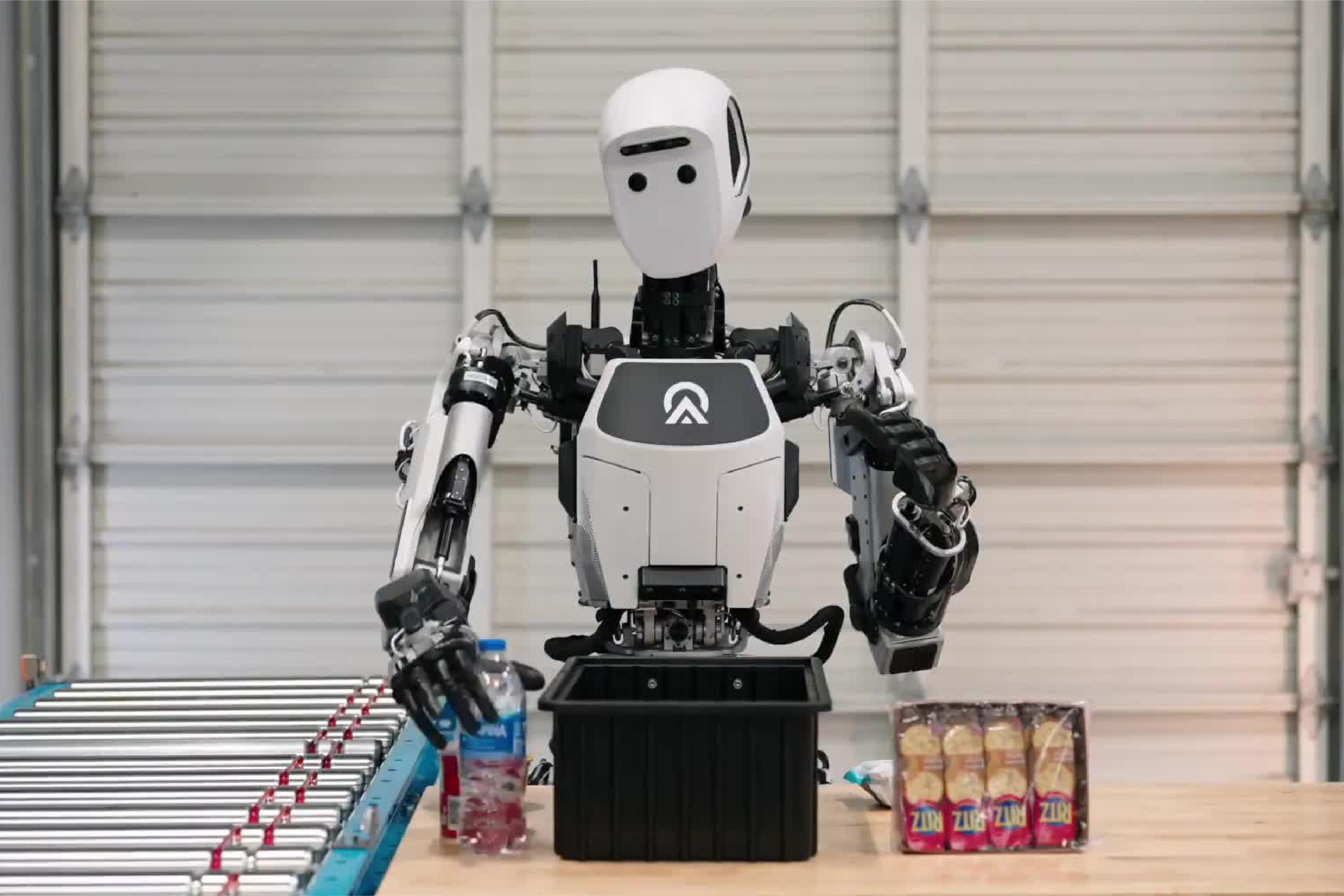 A humanoid robot works at a warehouse-style packing station, moving items between a table, bin and conveyor belt.