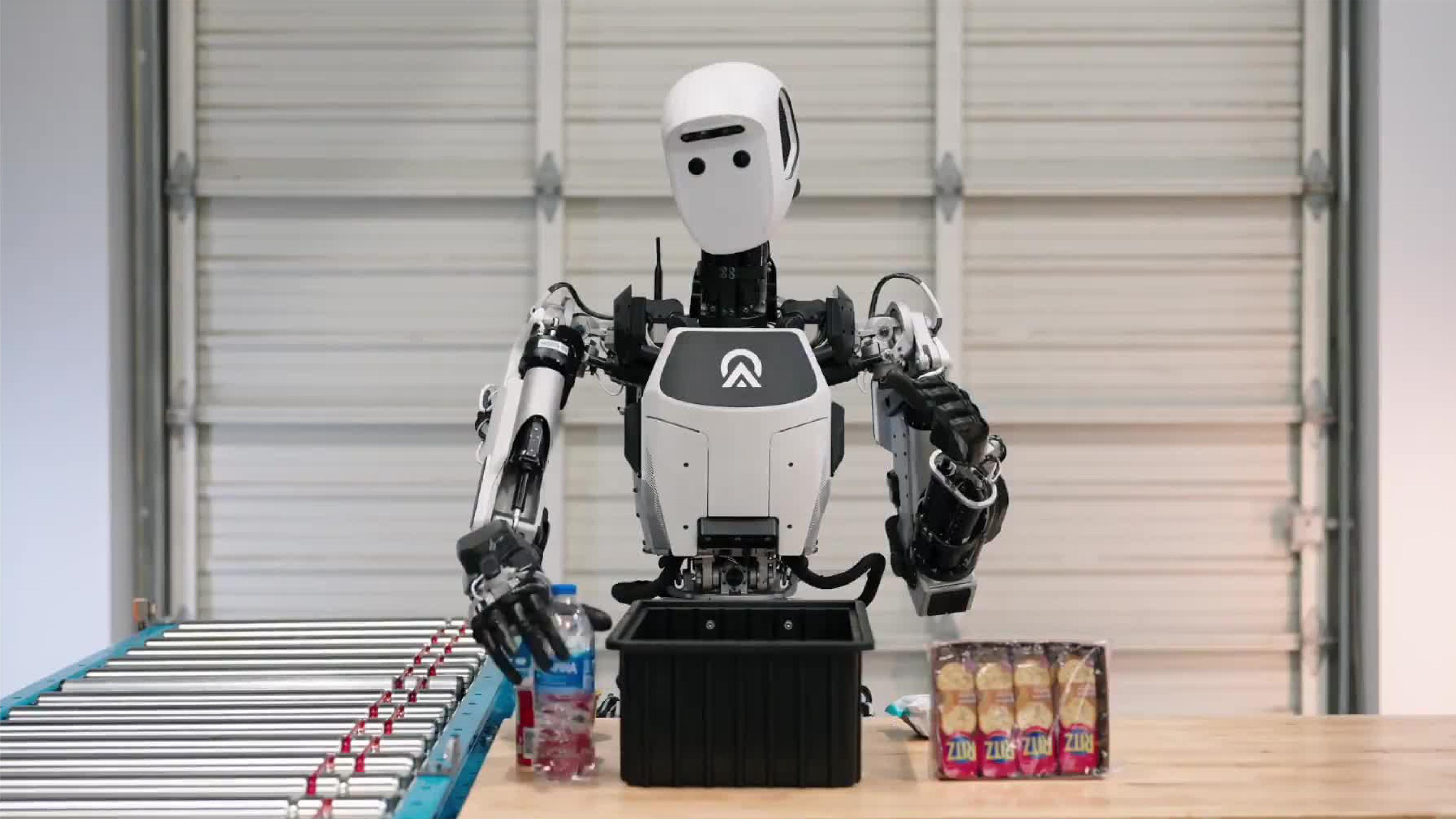 A humanoid robot works at a warehouse-style packing station, moving items between a table, bin and conveyor belt.